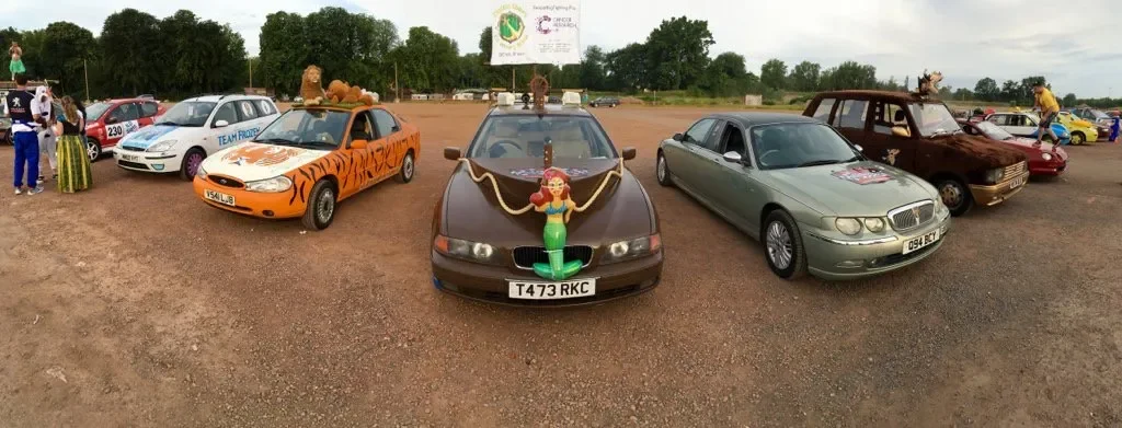 A vintage car decorated with a Hula girl figure in front, parked among other classic and customized cars at a car show on a dirt field.