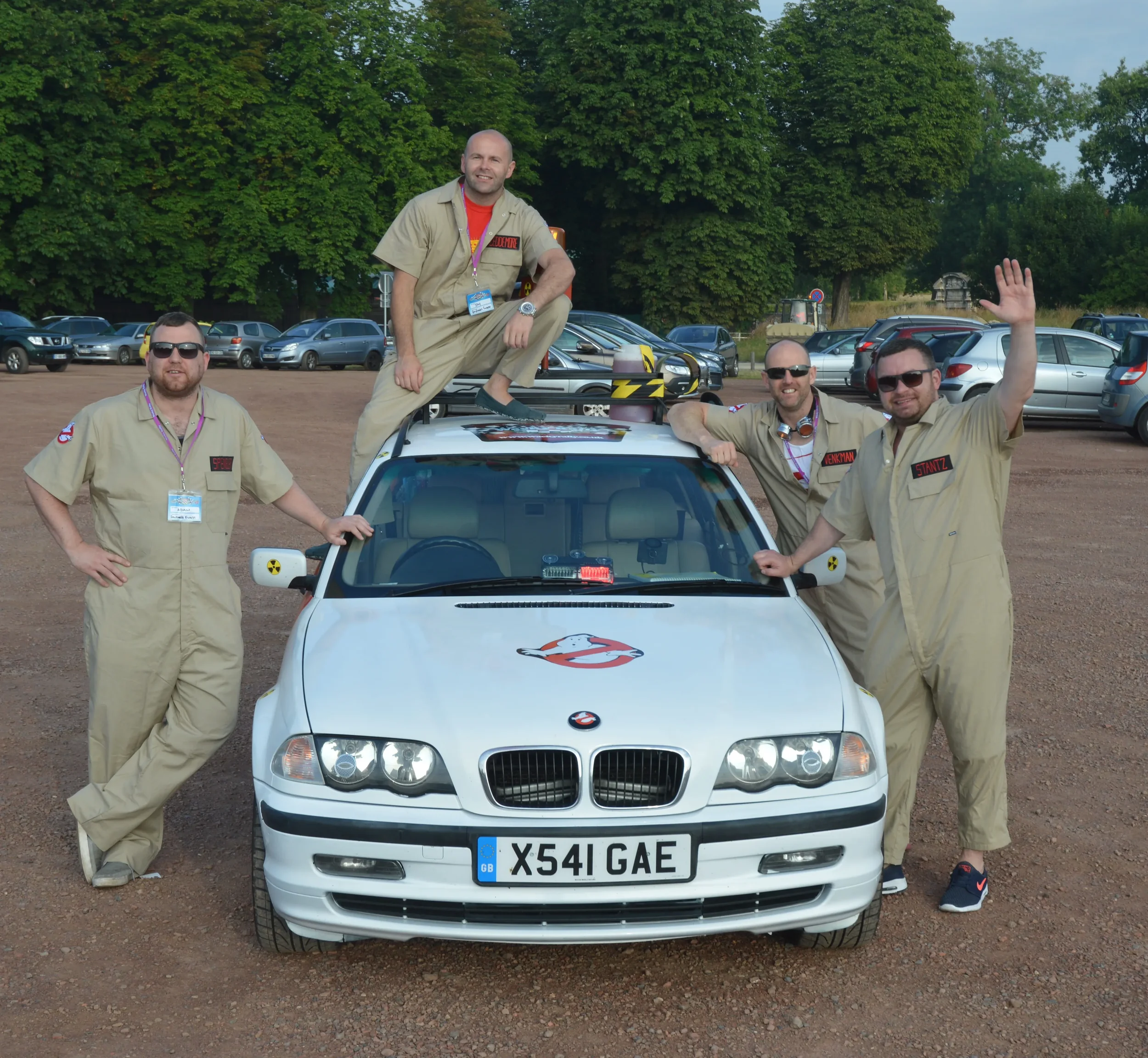 Four men dressed as ghostbusters pose around a white car with ghostbusters logo, in a parking lot with trees in the background.
