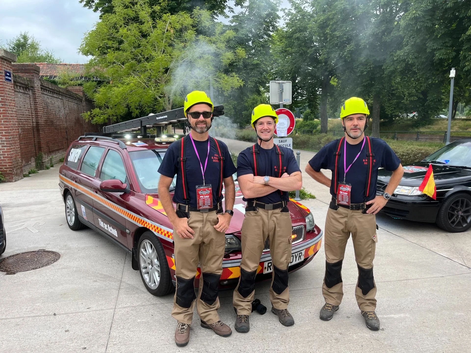 Three emergency responders standing in front of a red emergency vehicle, wearing yellow helmets and dark shirts, with trees and a brick wall in the background.