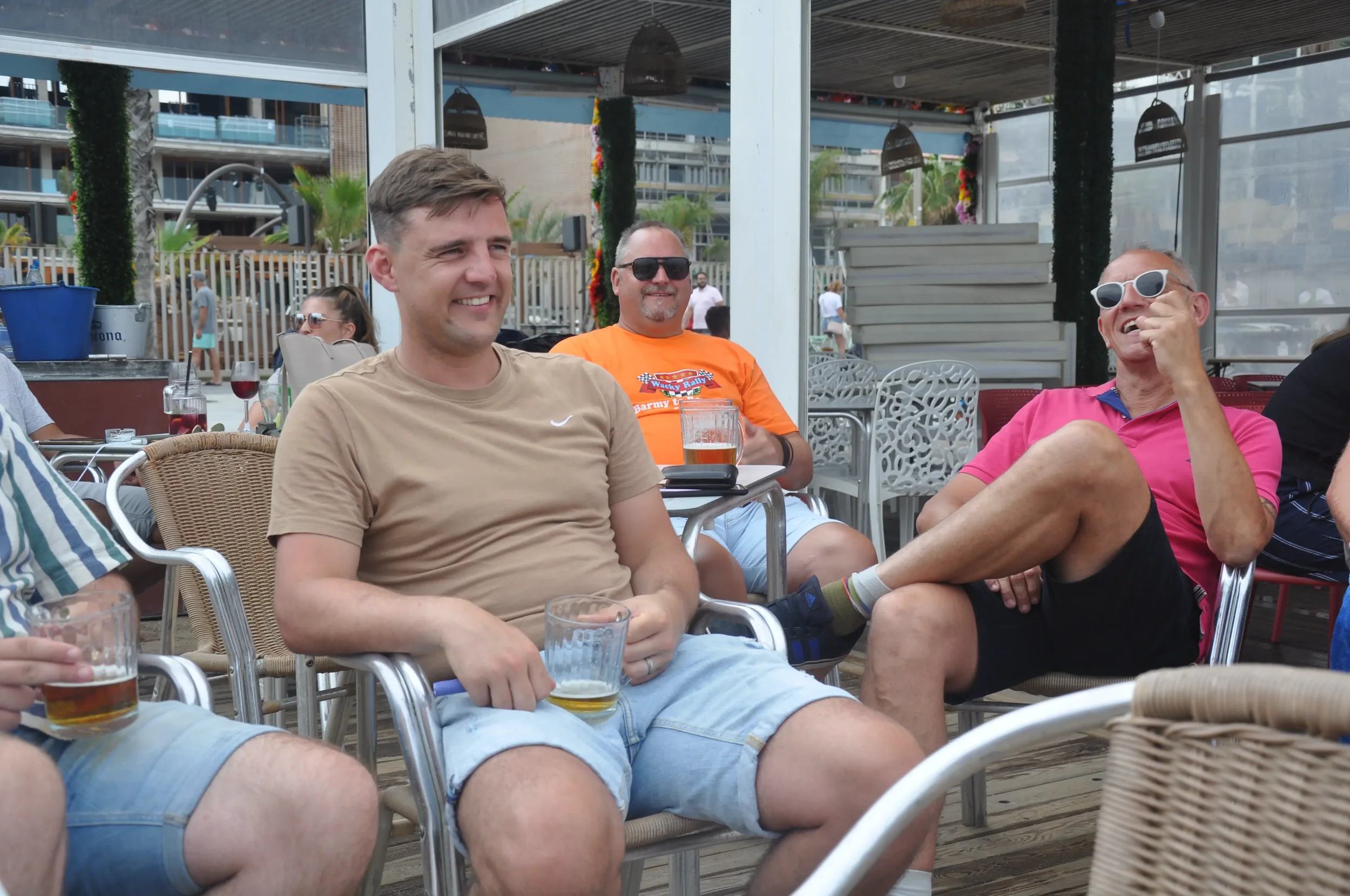 Group of friends sitting at an outdoor patio, smiling and laughing while holding drinks, with a man in orange shirt and sunglasses in the background.