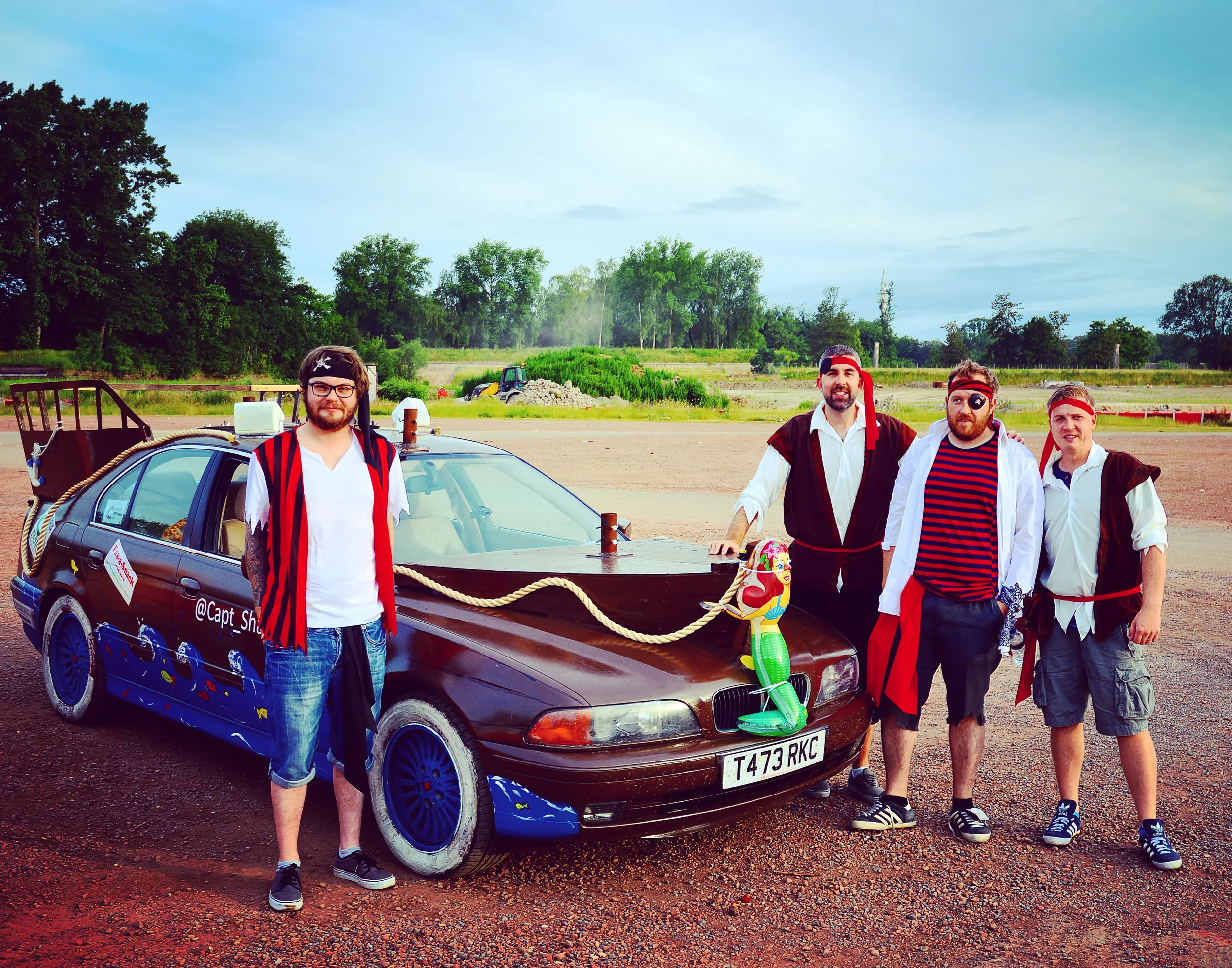 Four men dressed as pirates standing next to a decorated car with a film theme in an open area with trees and construction equipment in the background.