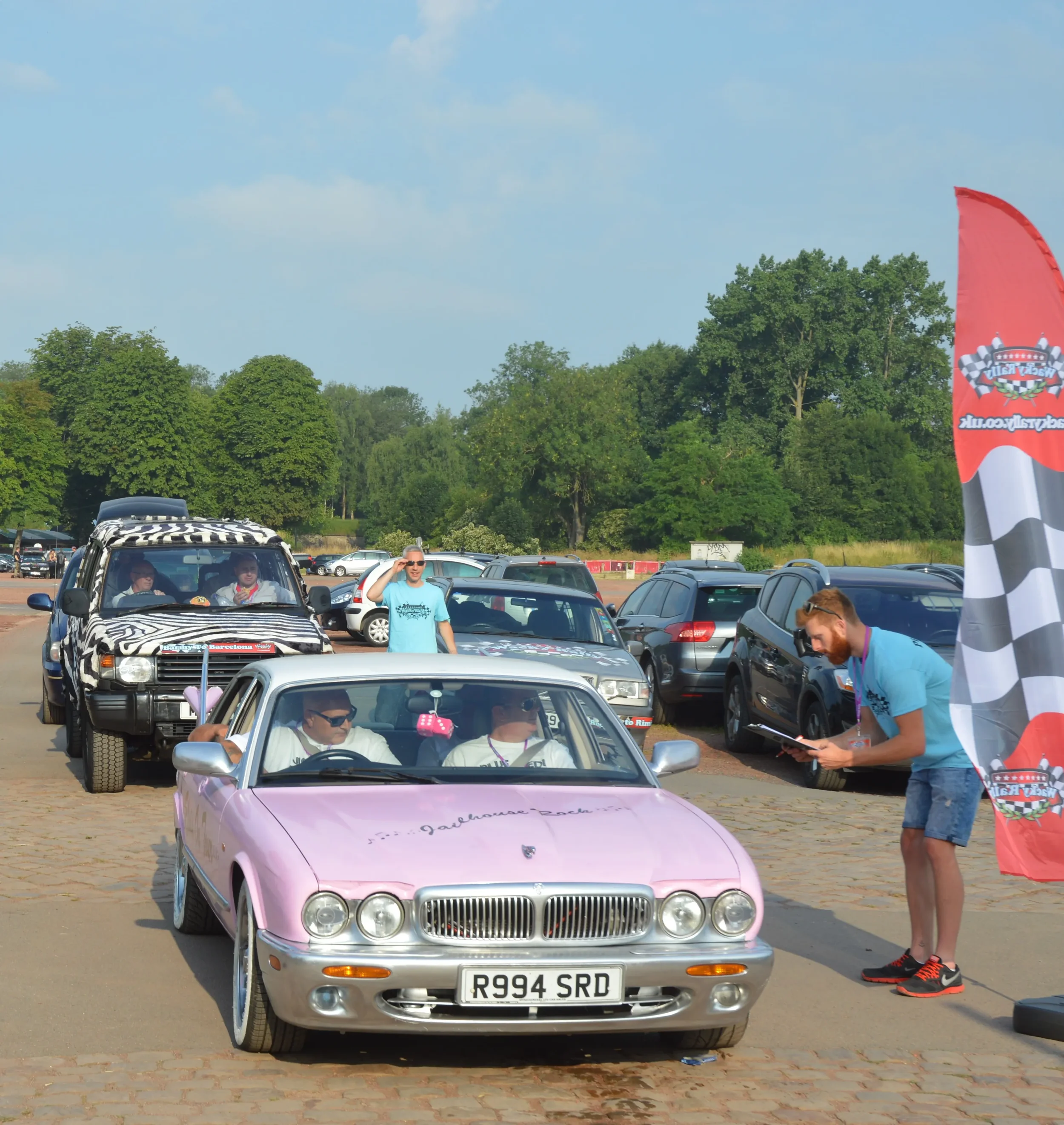 A pink classic convertible car with a UK license plate R994 SRD at the front, parked in a lot, with people sitting inside. A man is standing next to the car, wearing sunglasses and a white shirt, and another man is standing with a clipboard near a ta