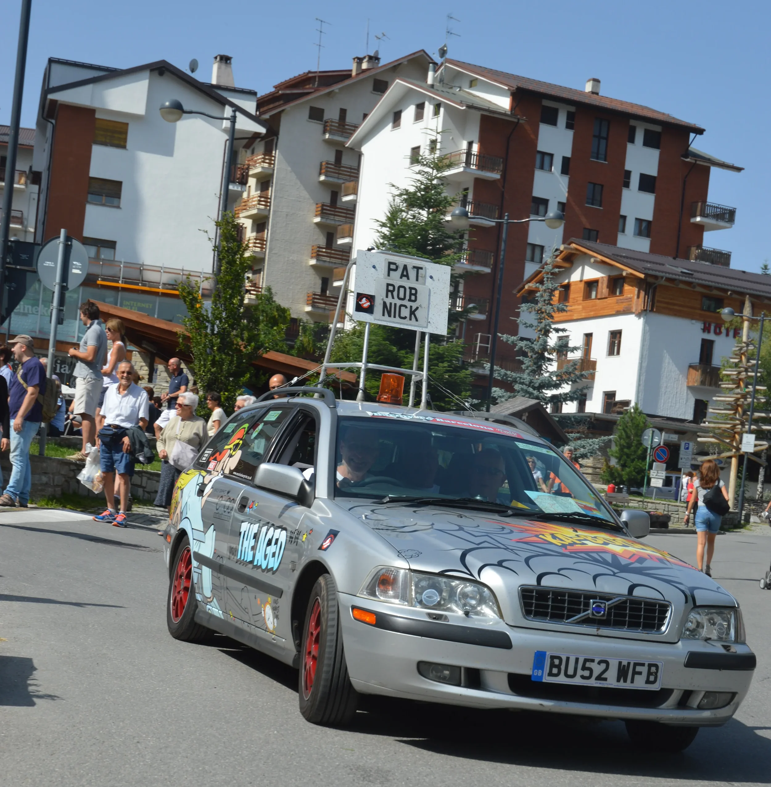 A silver Volvo car decorated with colorful artwork and the text "The Age" on the side, driving down a street with people walking nearby, in a town or city with multi-story buildings and hotels in the background.