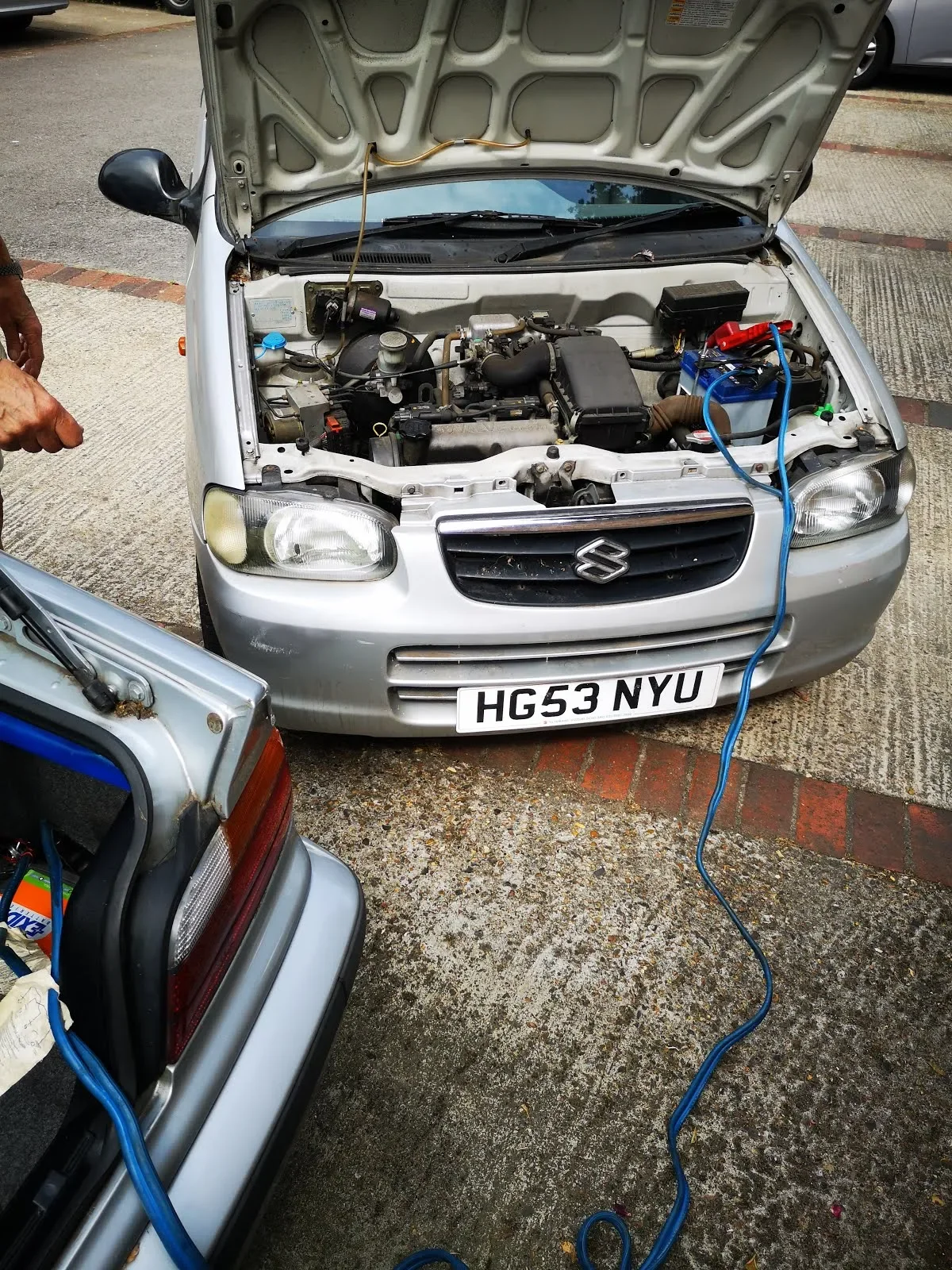Silver Suzuki car with open hood, connected to a roadside battery jump starter via blue jumper cable, on a concrete and brick road.