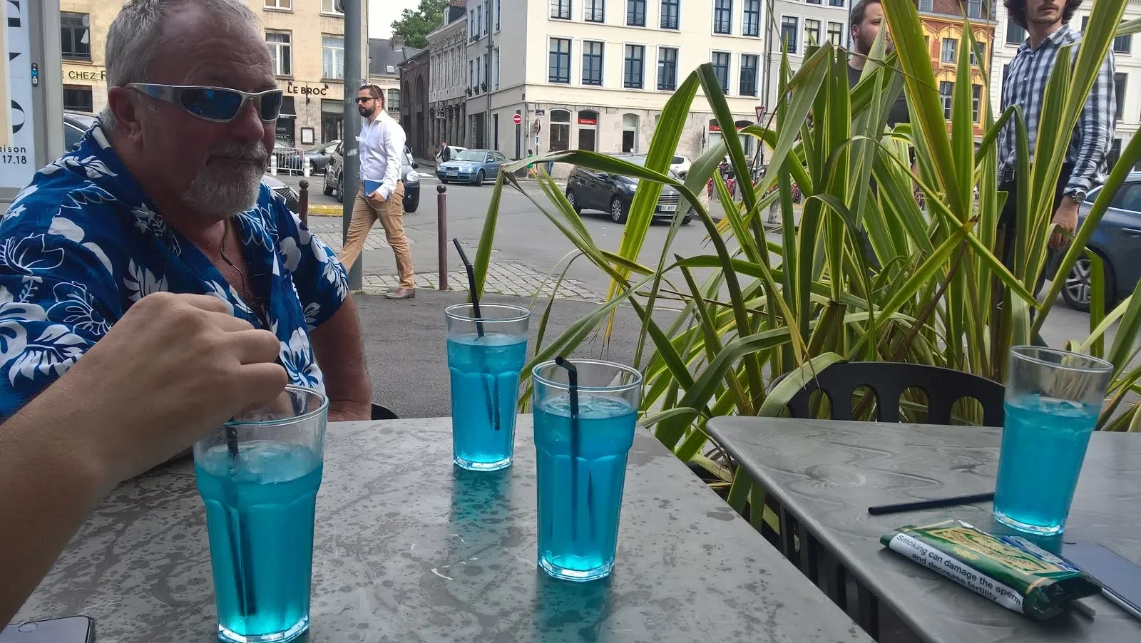 Man in a blue wave-pattern shirt with sunglasses sitting at an outdoor table with four blue drinks, with a city street scene and other people in the background.