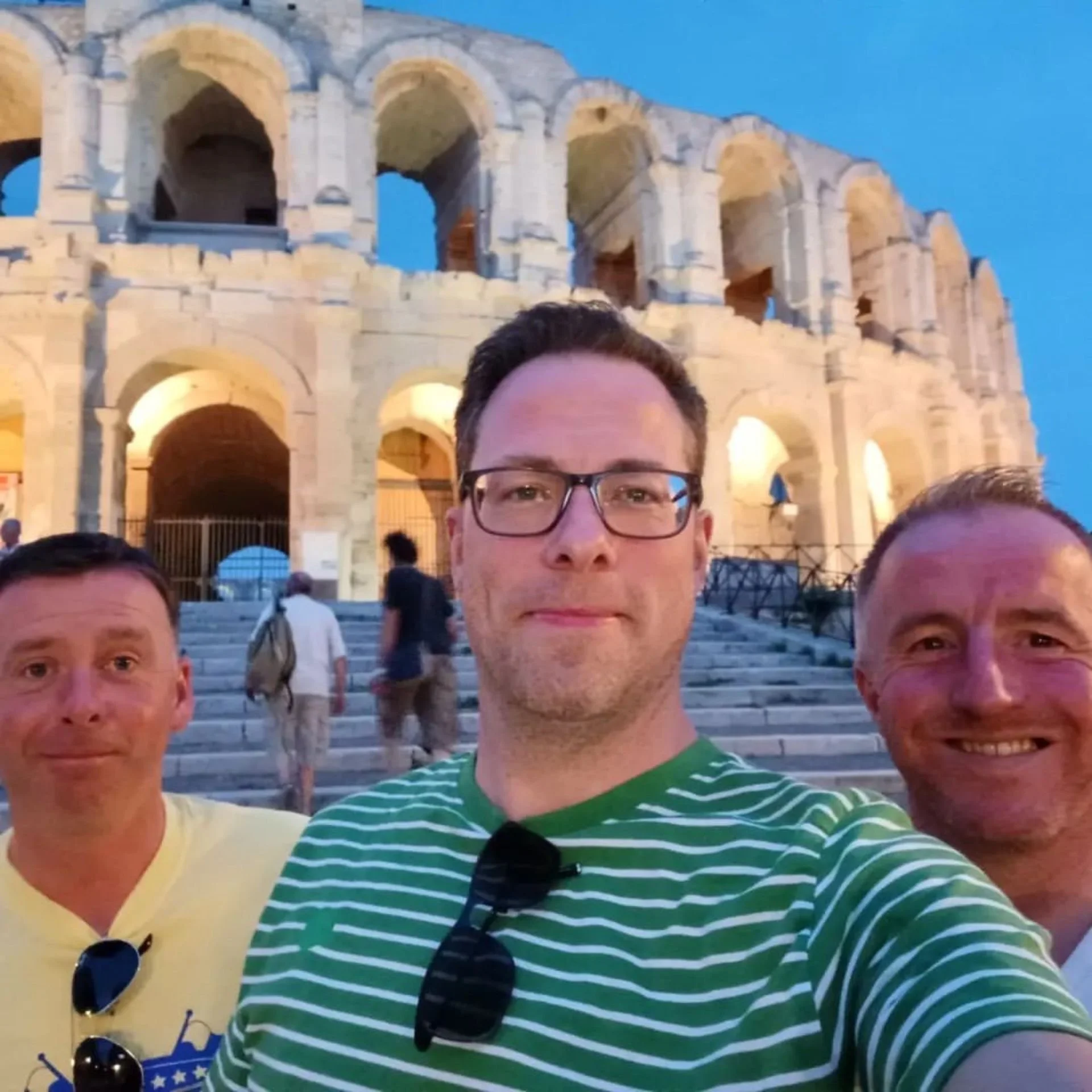 Three men taking a selfie in front of the Roman Colosseum during evening, with steps and other people in the background.