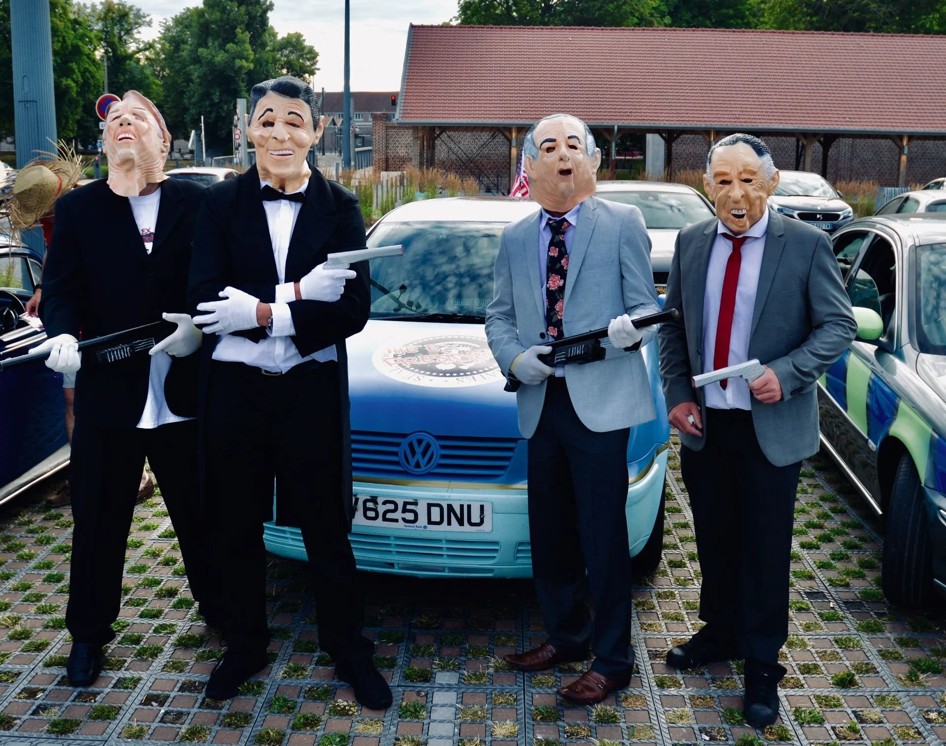 Four people wearing masks with exaggerated facial features, dressed in suits, standing in a parking lot with cars behind them. They are holding toy guns and papers, posing as a crime or mafia scene.