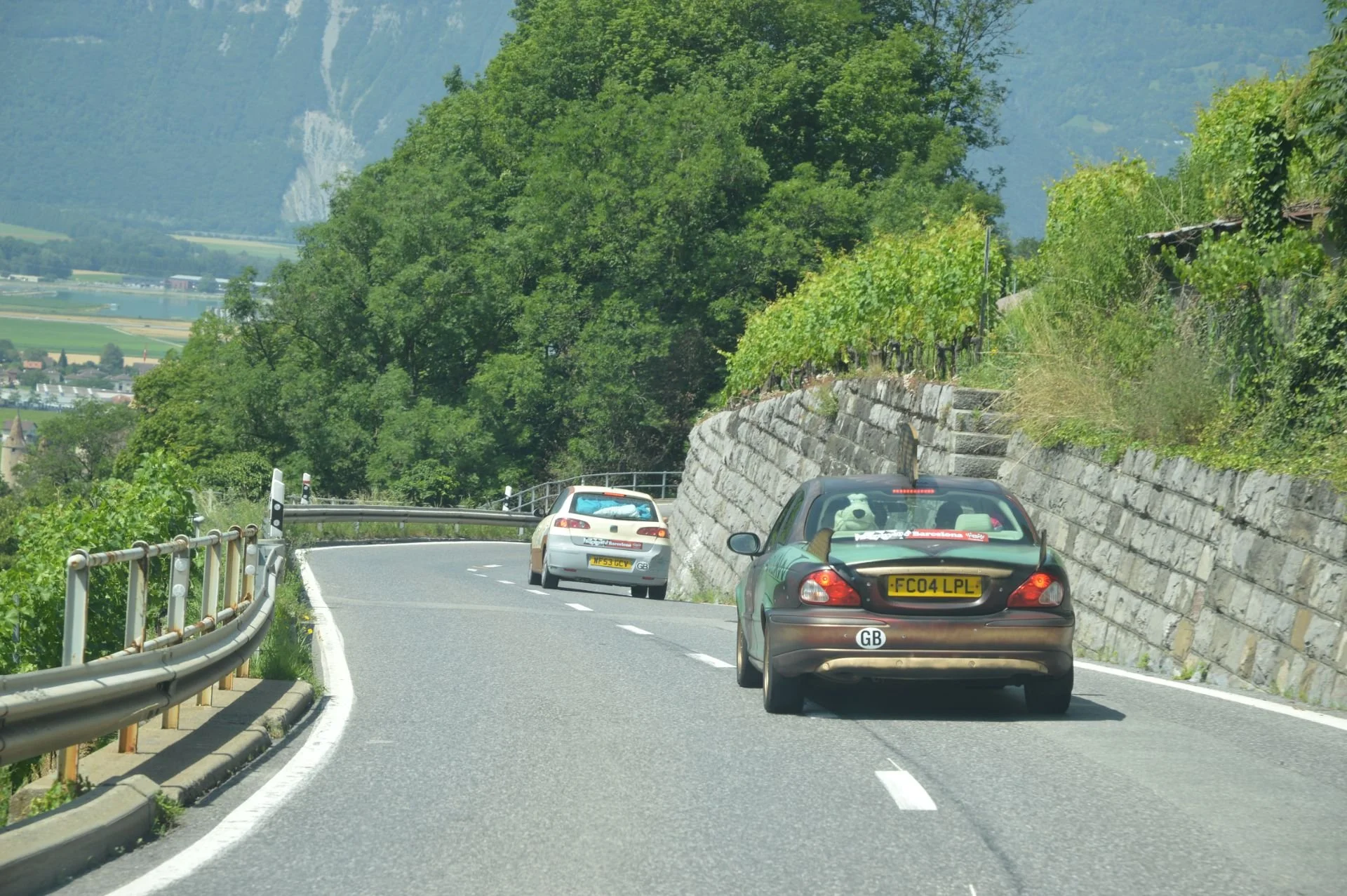 Two cars driving on a winding mountain road with guardrails on the left and a stone wall on the right, surrounded by green trees and hills in the background.
