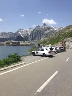 Parked cars on the side of a mountain road overlooking a body of water, mountains with snow-capped peaks in the background under a blue sky with clouds.