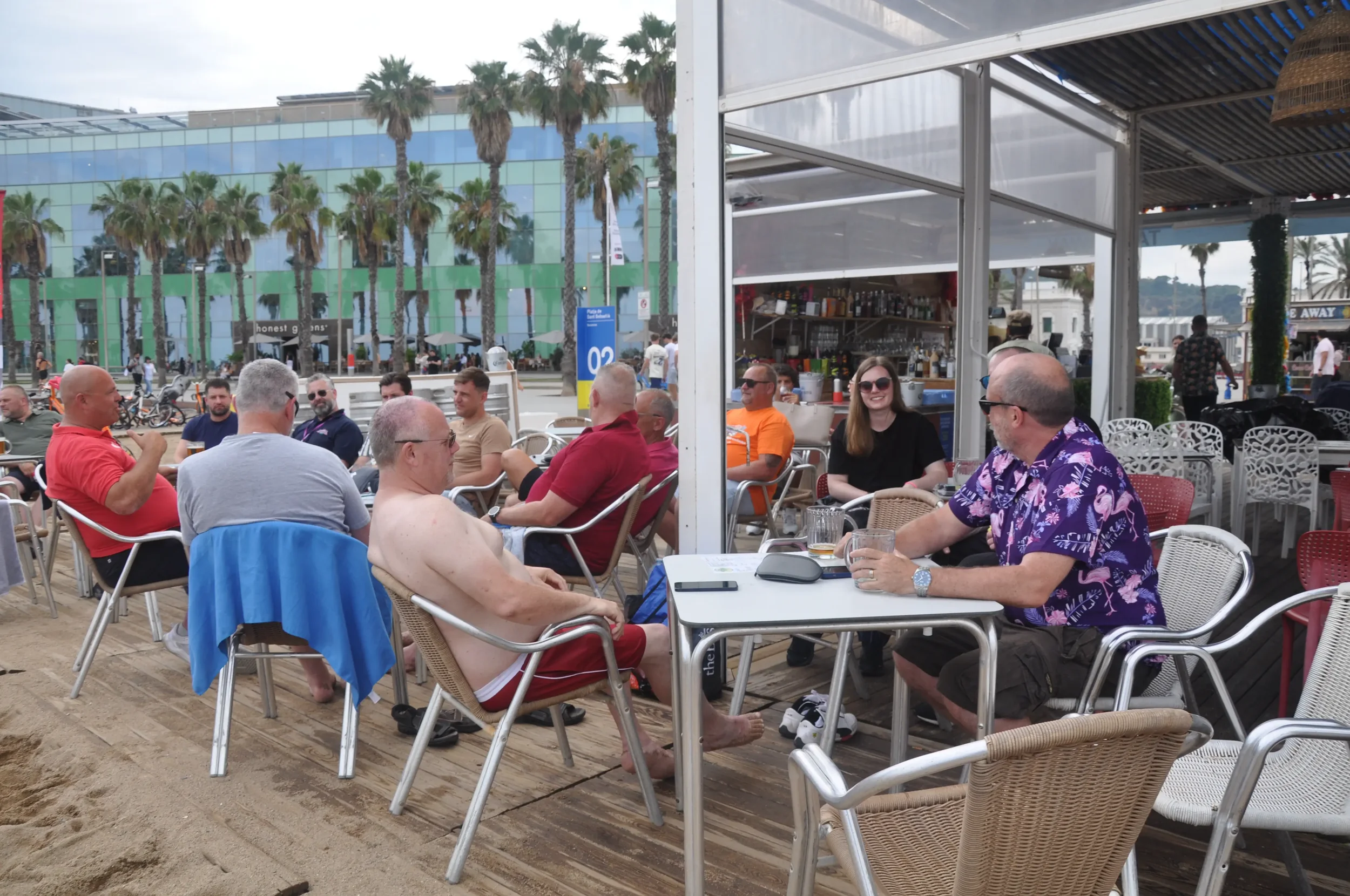 Group of people sitting and chatting at an outdoor bar or restaurant near the beach, with palm trees and a modern glass building in the background.
