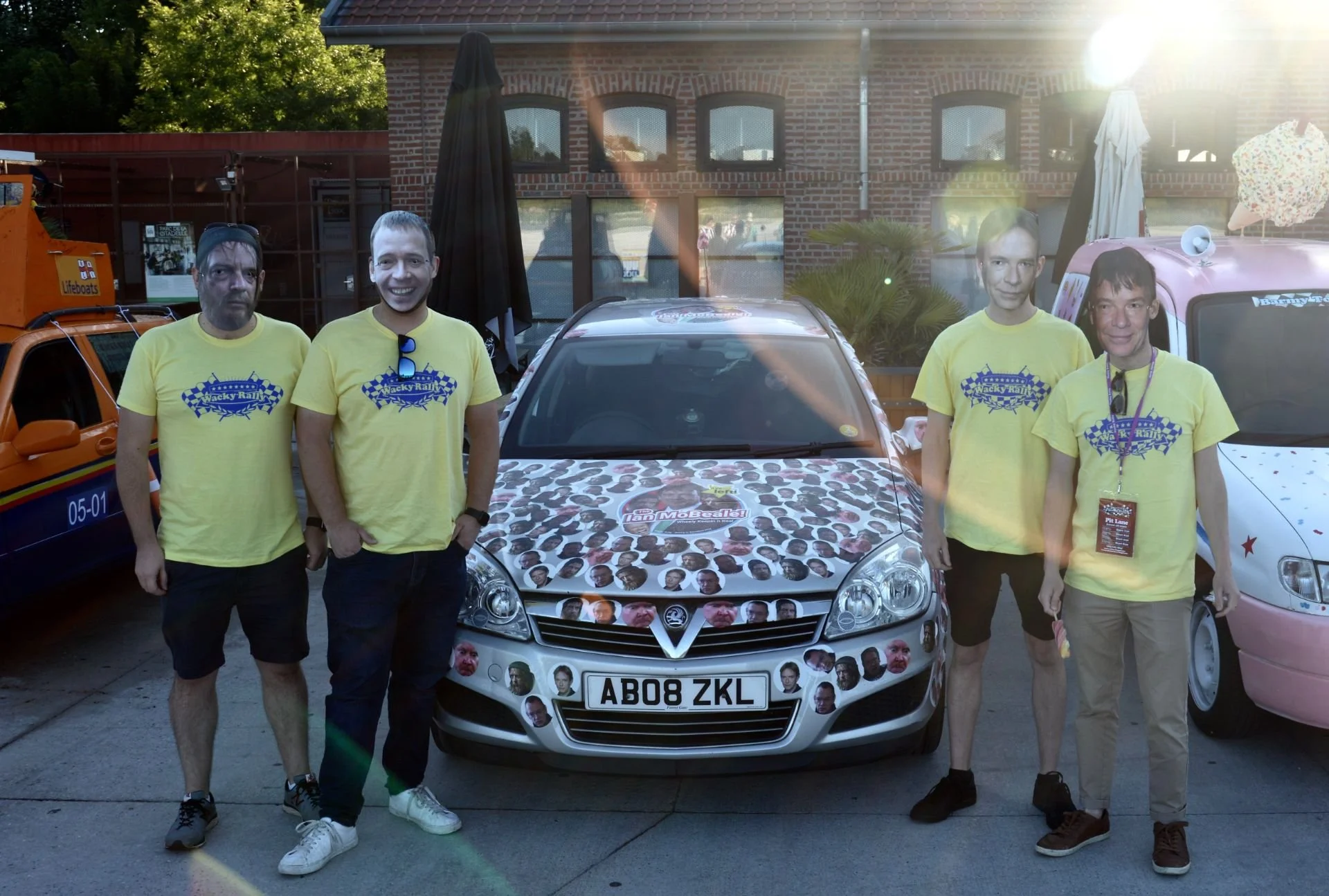 Four men standing outdoors in front of a decorated car, all wearing yellow shirts with a racing-themed logo, with a brick building and another decorated vehicle in the background.
