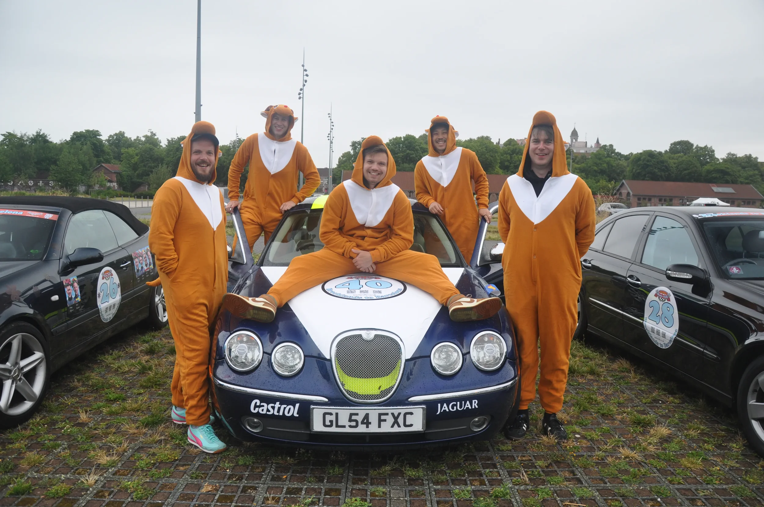 Group of six adults dressed in lion onesies around a vintage Jaguar race car at an outdoor event.