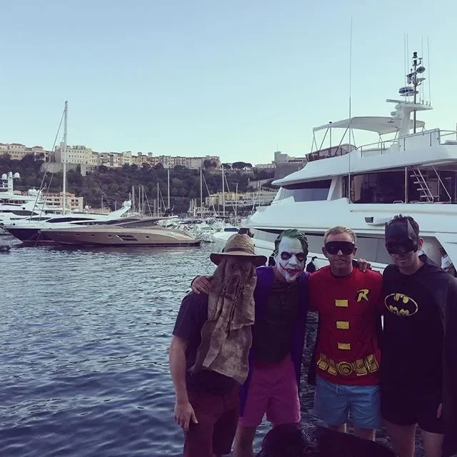 Four people dressed as Batman, Joker, Robin, and another character stand together near boats and yachts docked in a harbor with buildings in the background.