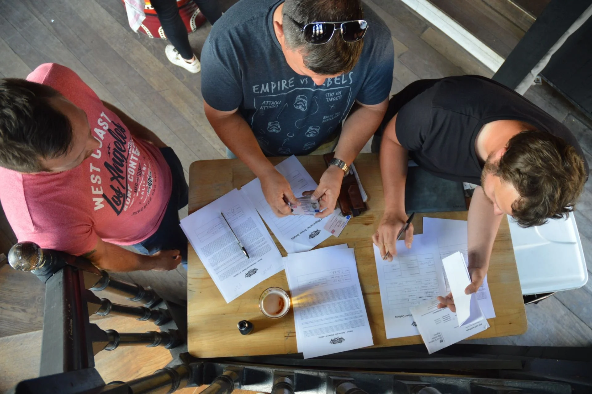 Three men are gathered around a wooden table with documents and a pen. One man is wearing sunglasses and a blue T-shirt, another in a black T-shirt, and the third in a pink T-shirt. A beer glass is on the table, and the setting appears to be indoors 