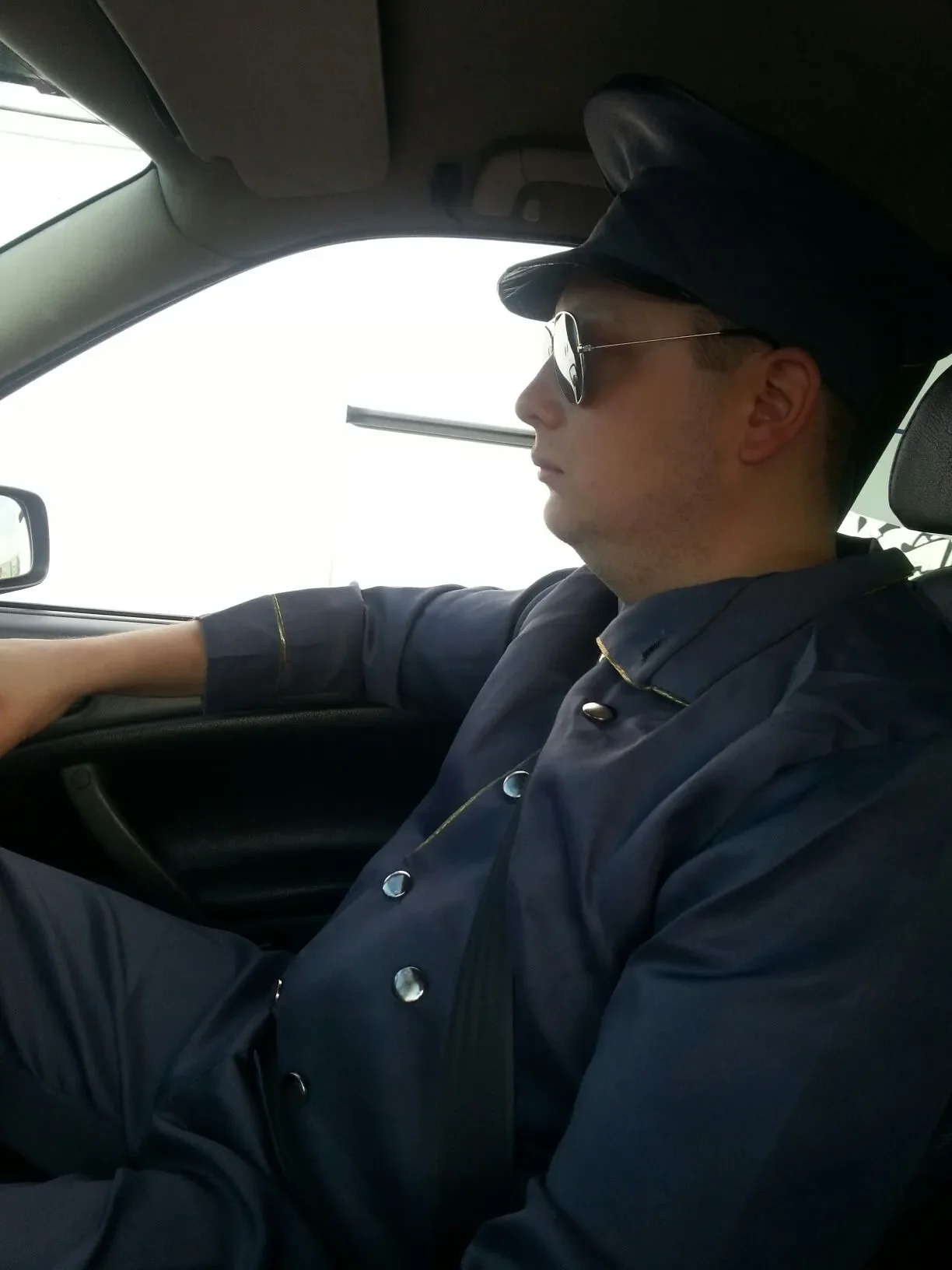 A man dressed as a police officer wearing a dark blue uniform, hat, and sunglasses sitting in the driver's seat of a car.