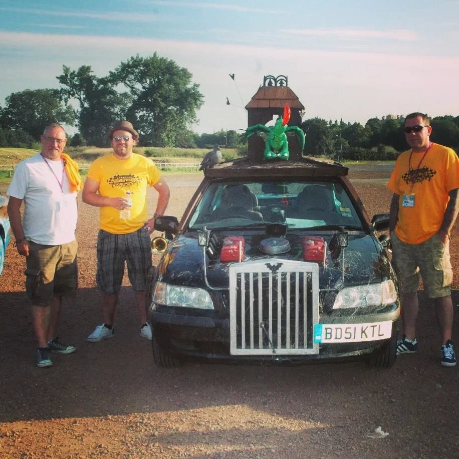 Three men standing next to a black car with a large dragon puppet on top, outdoors on a sunny day.