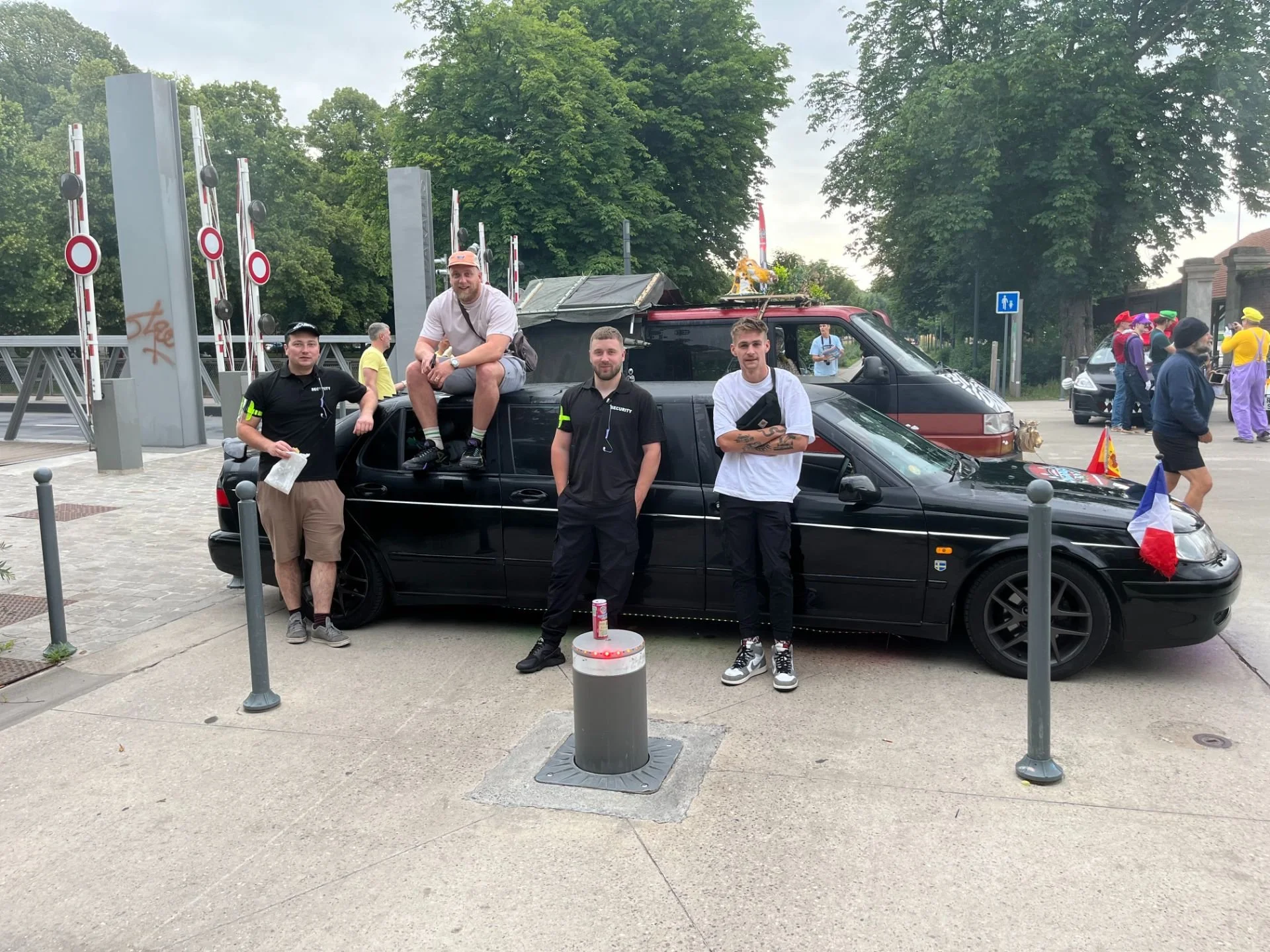Four men are standing and sitting around a black car with French flags attached. They are outdoors near a crossing gate and trees, with other people in the background, some dressed in colorful clothing.