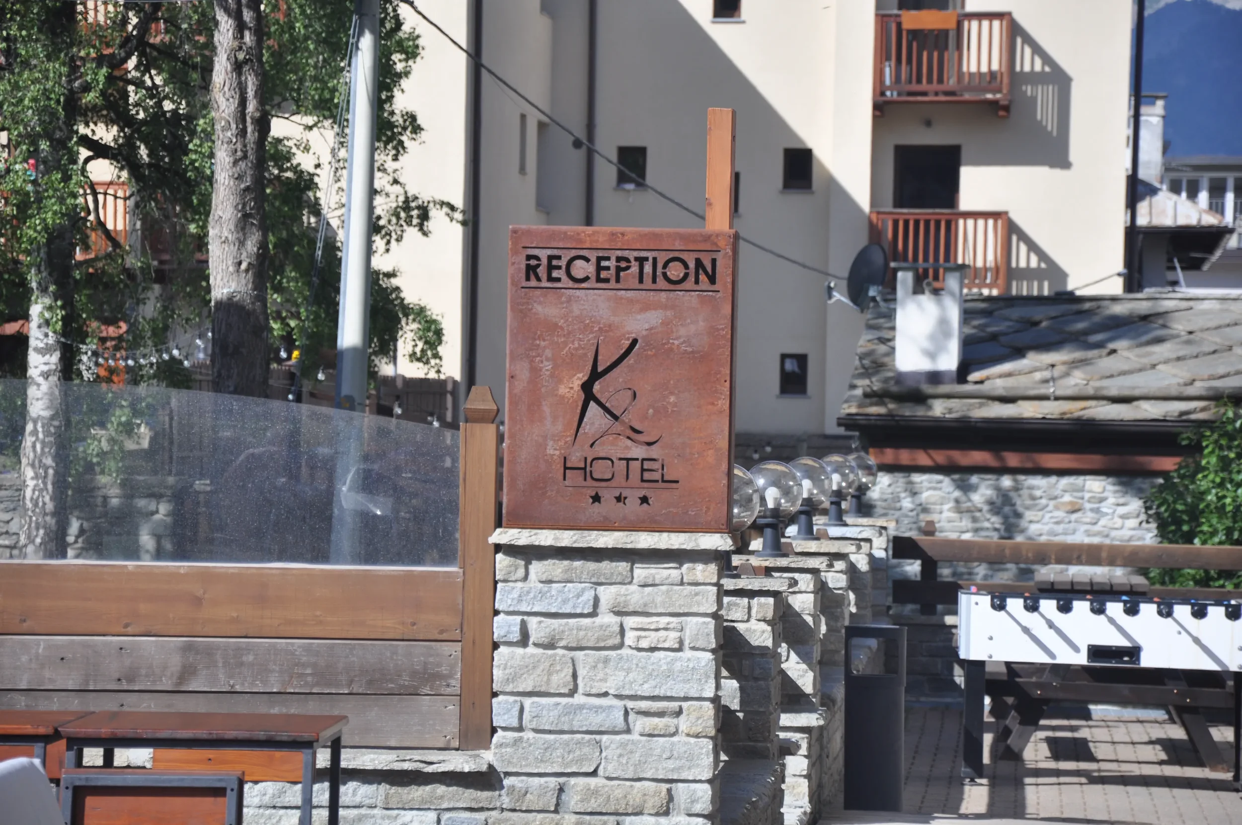 Hotel reception sign in front of a stone wall, with a logo, three stars, and a wooden patio area with a bench and a foosball table.