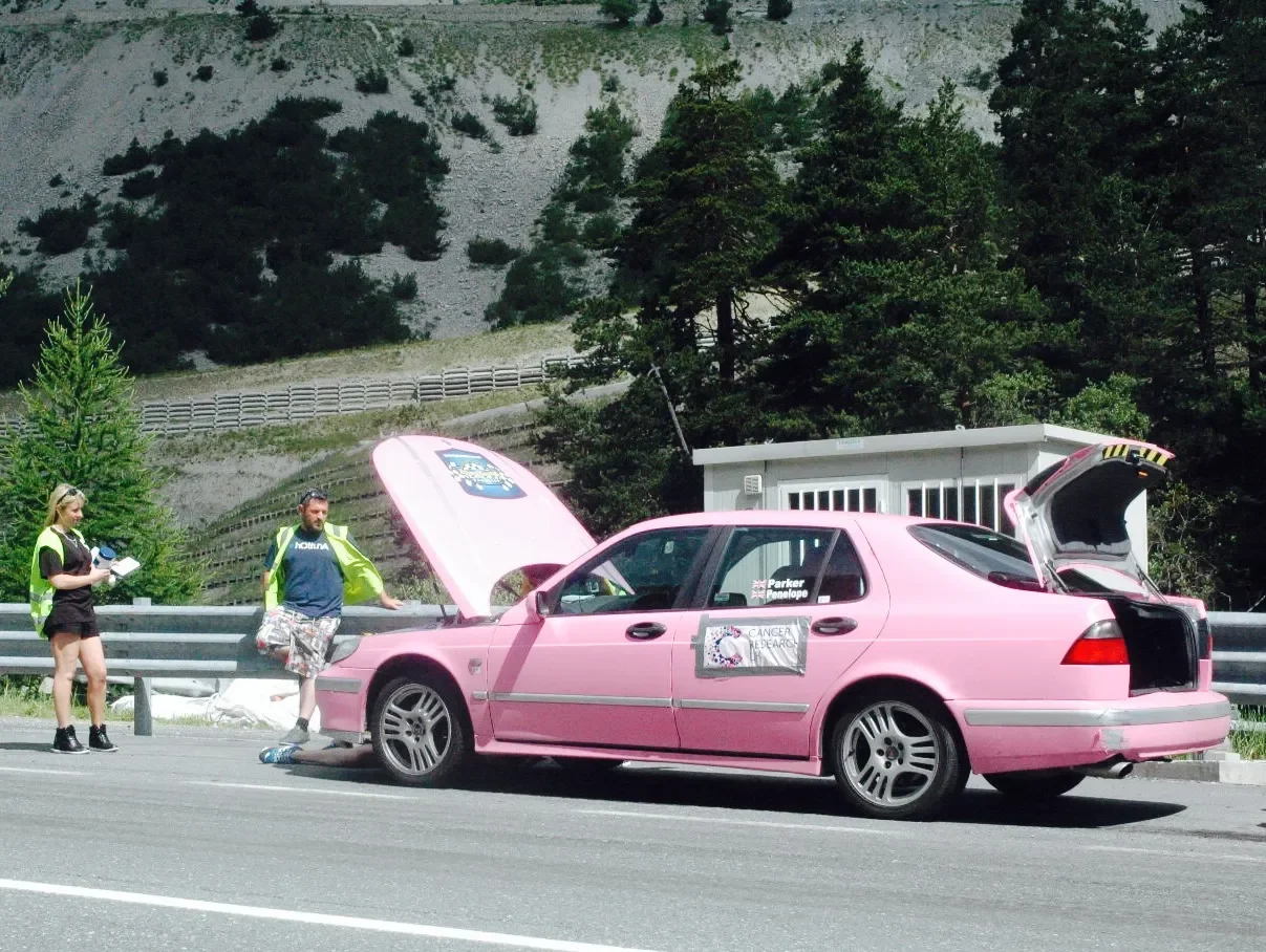 A pink car with open hood and trunk stopped on a highway. Two people, one woman and one man, are standing beside the car. The woman is taking notes, and the man is leaning on the car. There is a sign on the car door related to cancer research. Trees 