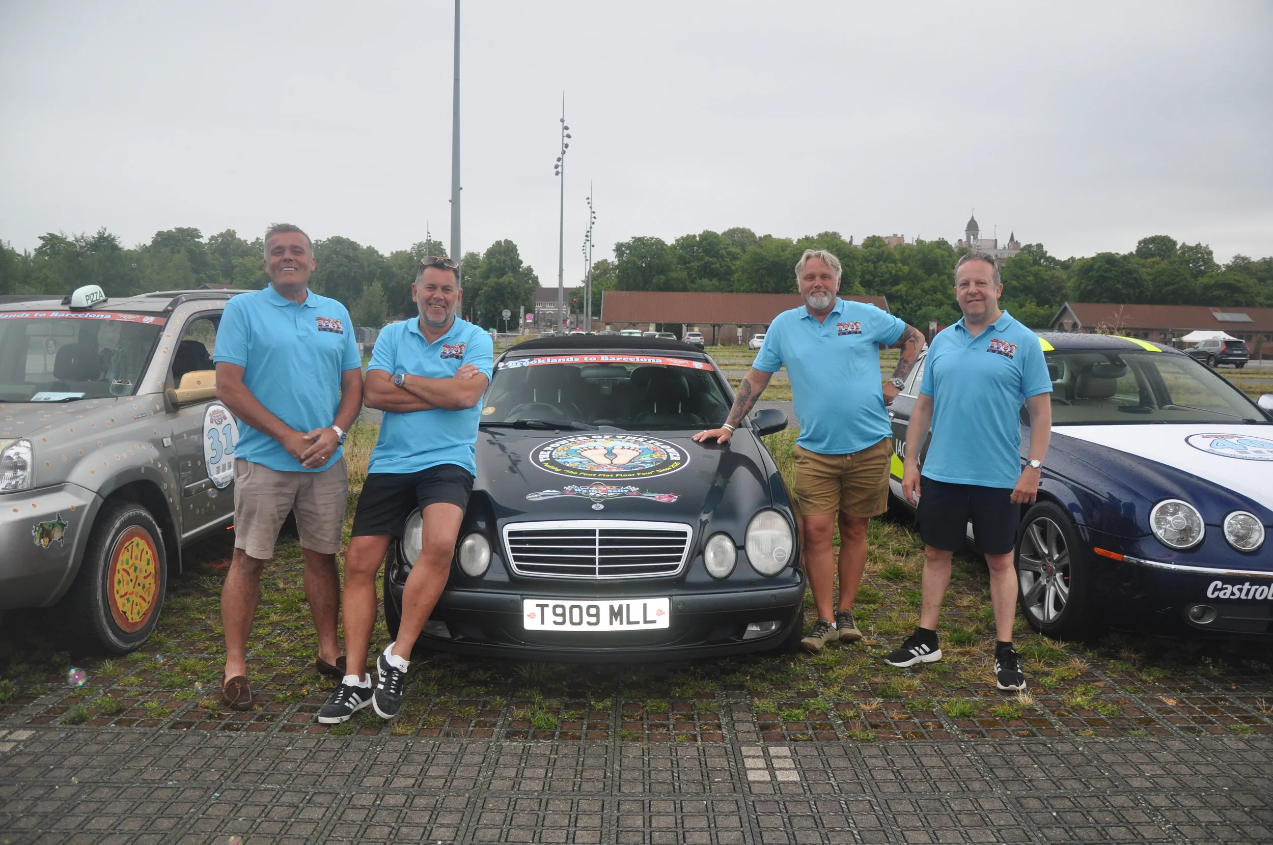 Five men standing outdoors near a row of cars, all wearing matching blue t-shirts. The cars are decorated with racing or event decals and are parked on a cobblestone surface. The background shows trees and a cloudy sky.