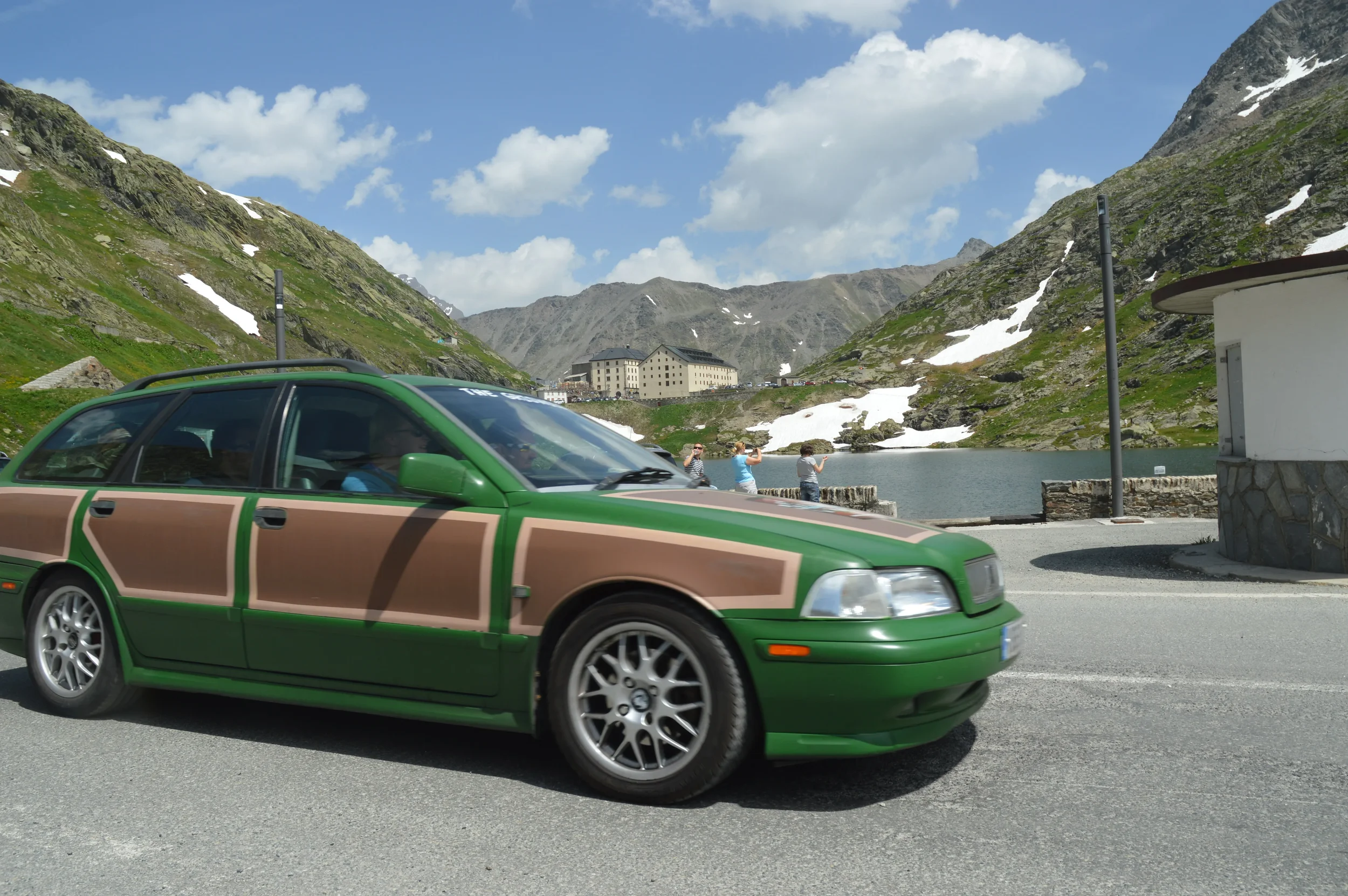 A green car with camouflage paint parked on a mountain road near a lake, with mountains, snow patches, and a building in the background under a partly cloudy sky.