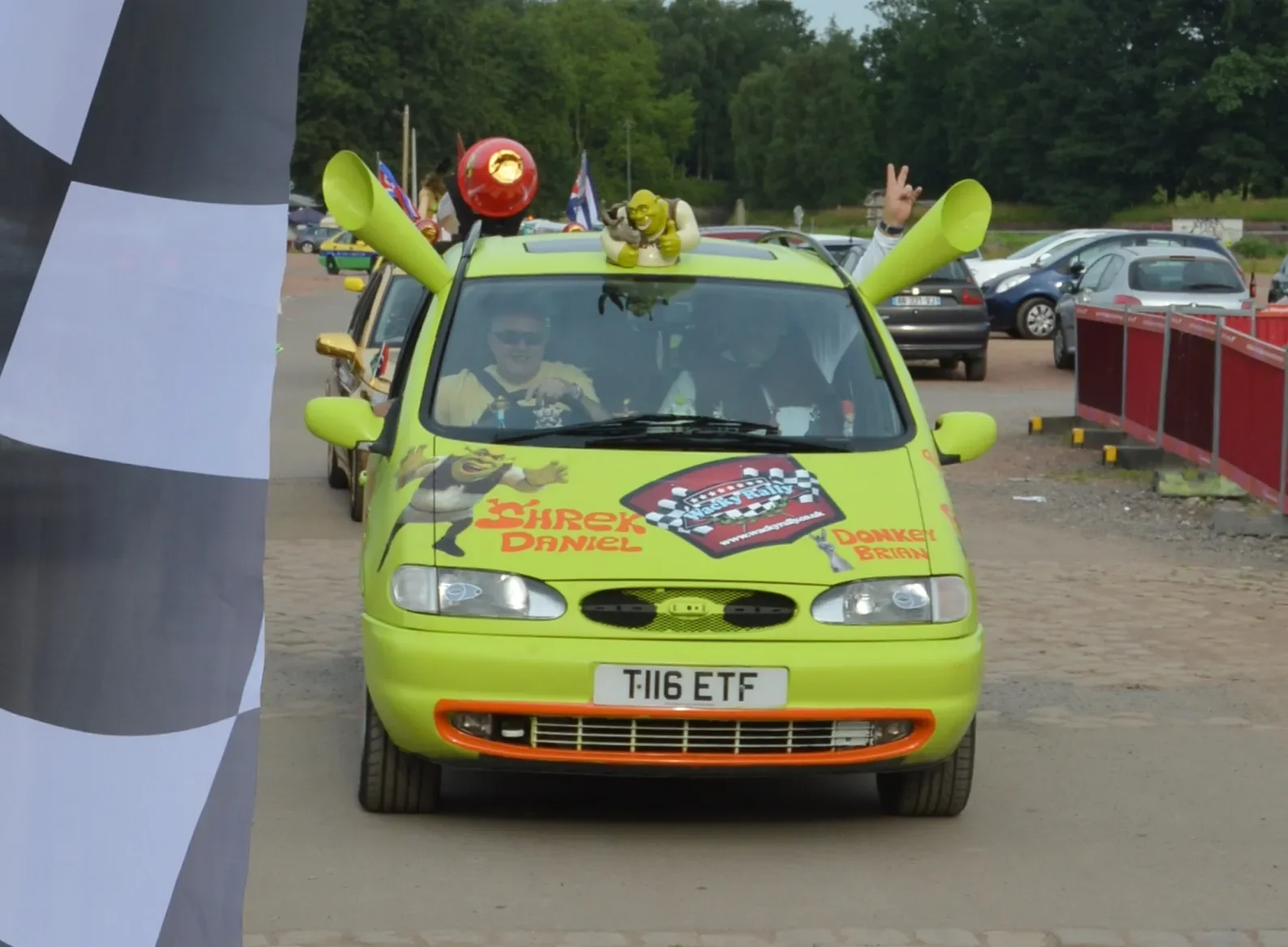 A bright green car decorated with characters from the animated movie Shrek, including Shrek, Donkey, and Fiona, participating in a race or parade with flags and decorations.