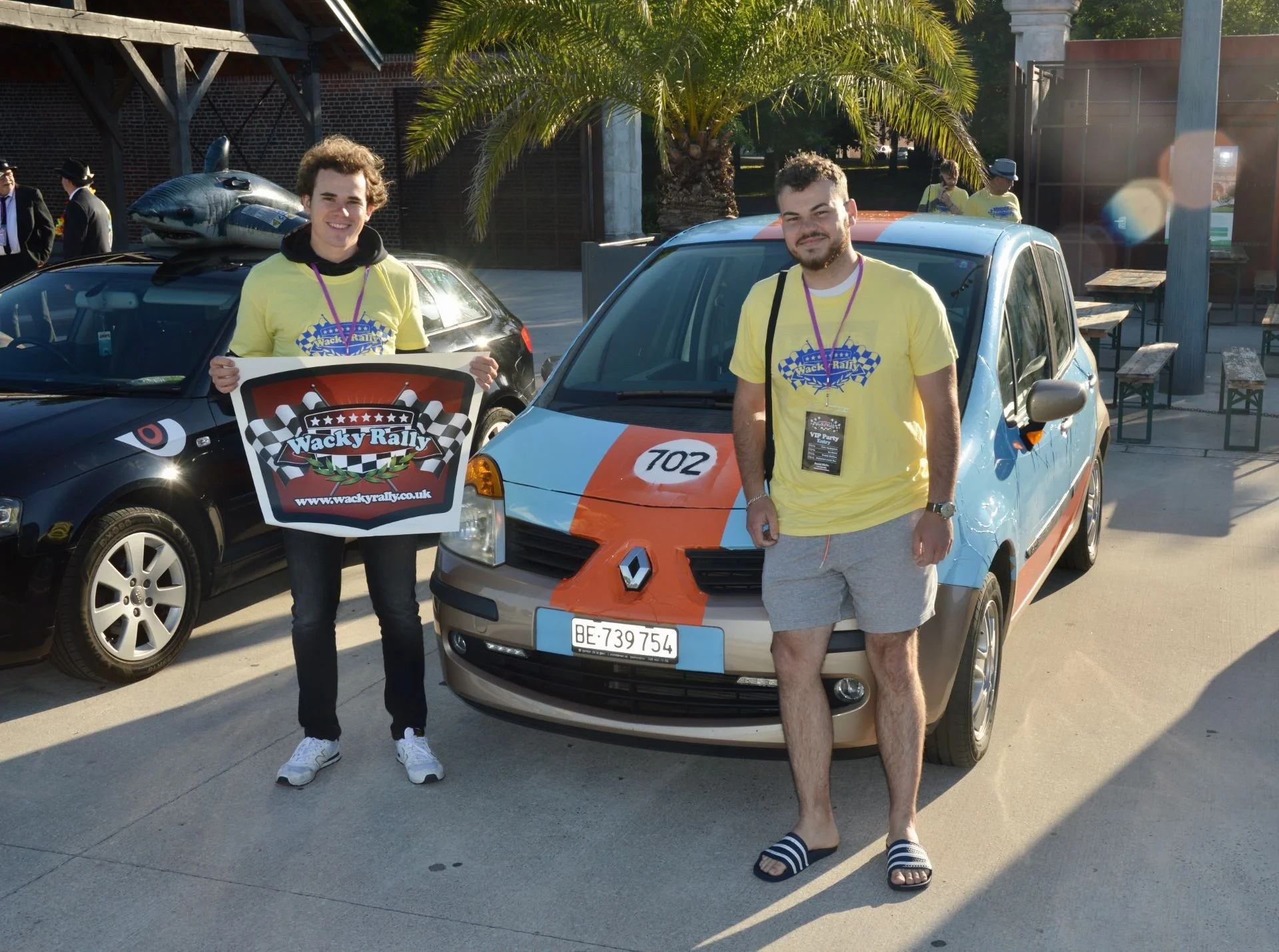 Two men standing in front of a blue Renault car with racing stripes, at a rally event. One man is holding a Wacky Rally sign, and both are wearing yellow T-shirts with rally-themed logos. There are other cars and people in the background, under sunny