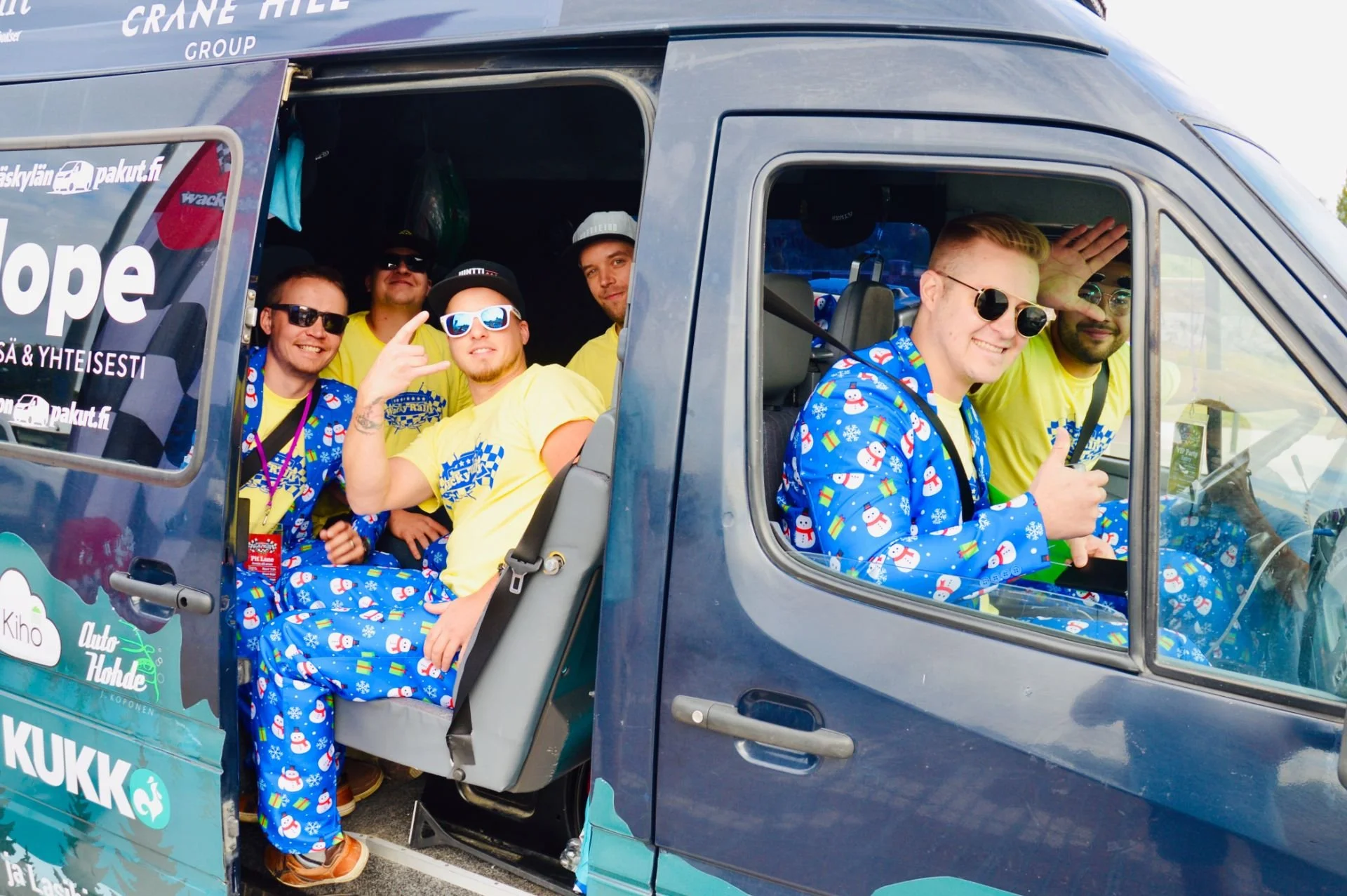 Group of seven young men sitting in a bus, wearing casual clothes with some in matching blue snowman-themed pajamas, some wearing sunglasses, smiling, and posing for the camera inside a festive vehicle decorated with logos and stickers.