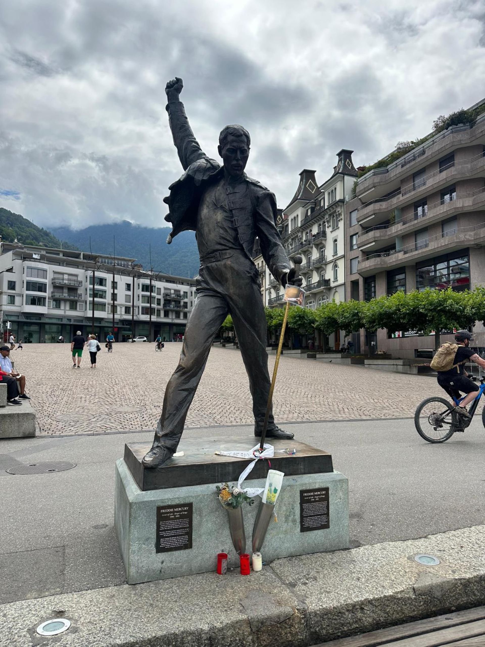 A statue of Freddie Mercury holding a microphone stand with a white ribbon and flowers, located in a public square with modern buildings, people, and mountains in the background.