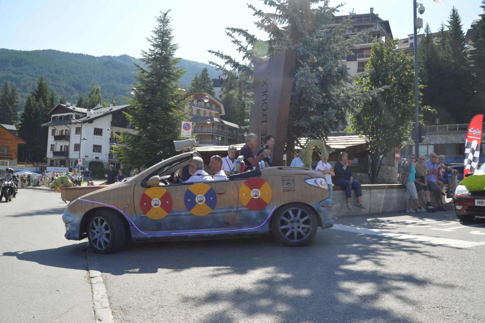 A convertible car decorated with beach-themed paint and foam swords on the side, carrying people, driving through a street with spectators and a mountainous background.