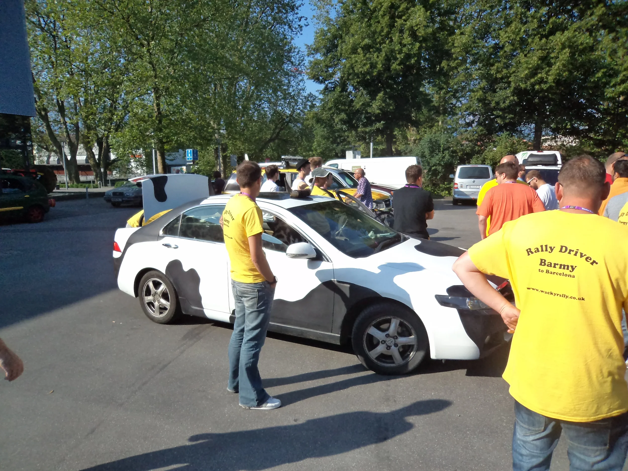 Group of rally car drivers and spectators gathered around a white rally car with a black hood, some wearing yellow shirts with text, in a parking lot on a sunny day.