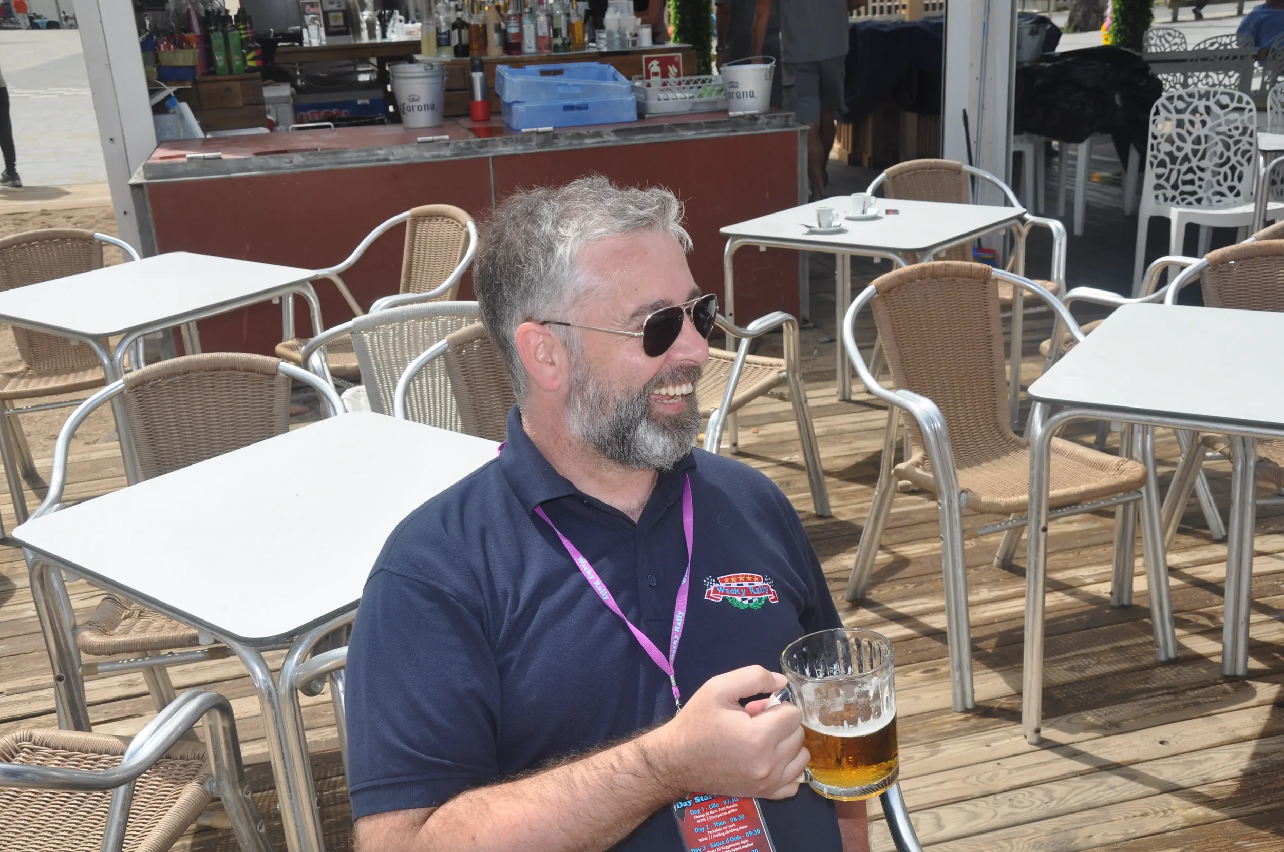 A smiling middle-aged man with gray hair and beard, wearing sunglasses and a navy blue shirt, sitting outdoors on a wooden deck with empty tables around him, holding a beer mug in his right hand. He appears to be enjoying a sunny day at a casual outd