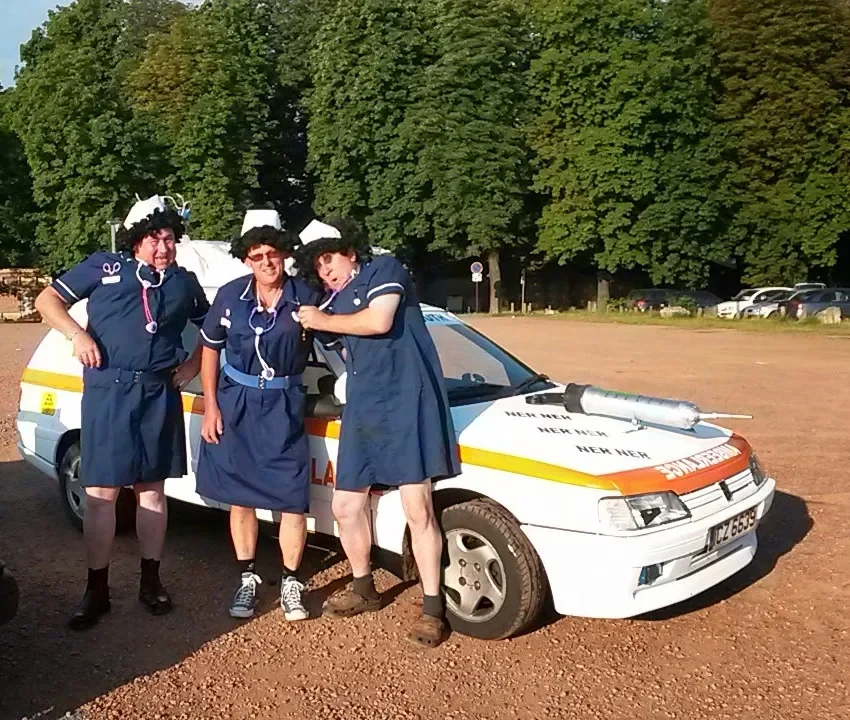 Three people dressed as nurses in costume stand in front of a vehicle on a dirt lot, with trees in the background.