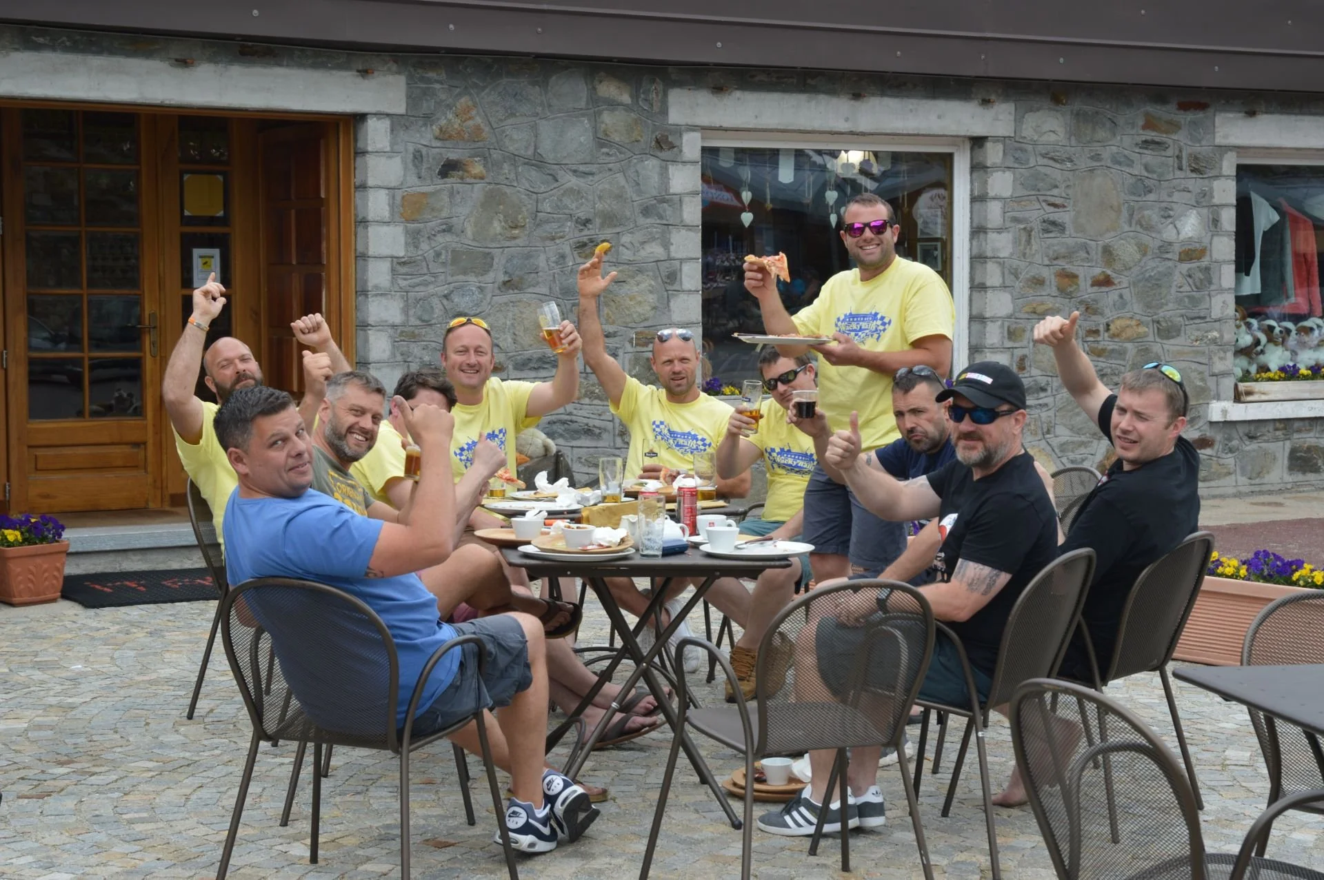 Group of men enjoying a meal and drinks outside a restaurant, some raising glasses and a man standing on a chair holding a slice of pizza.