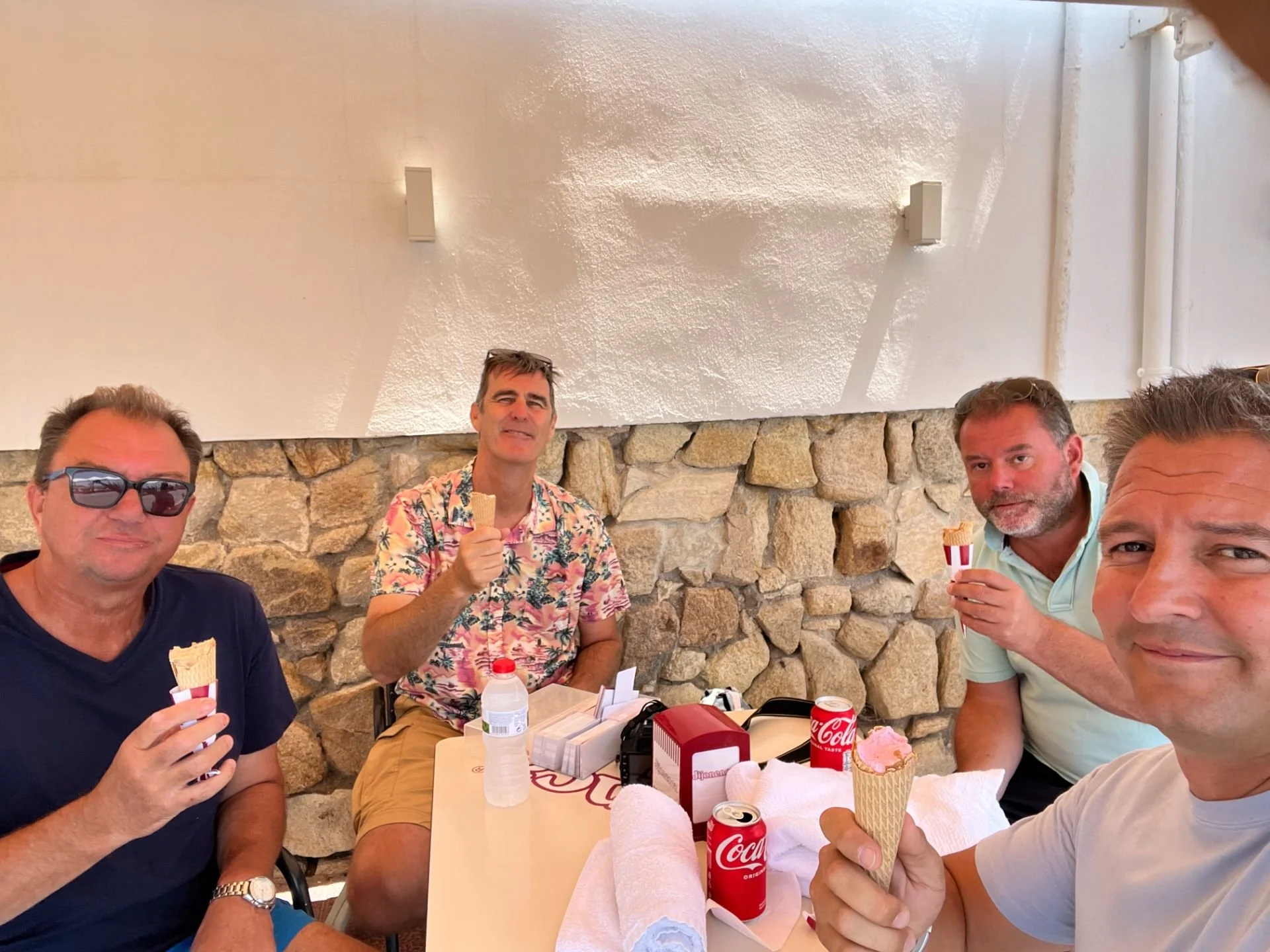 Four men sitting at a table, enjoying ice cream cones and drinks, inside a room with stone and white wall decor.