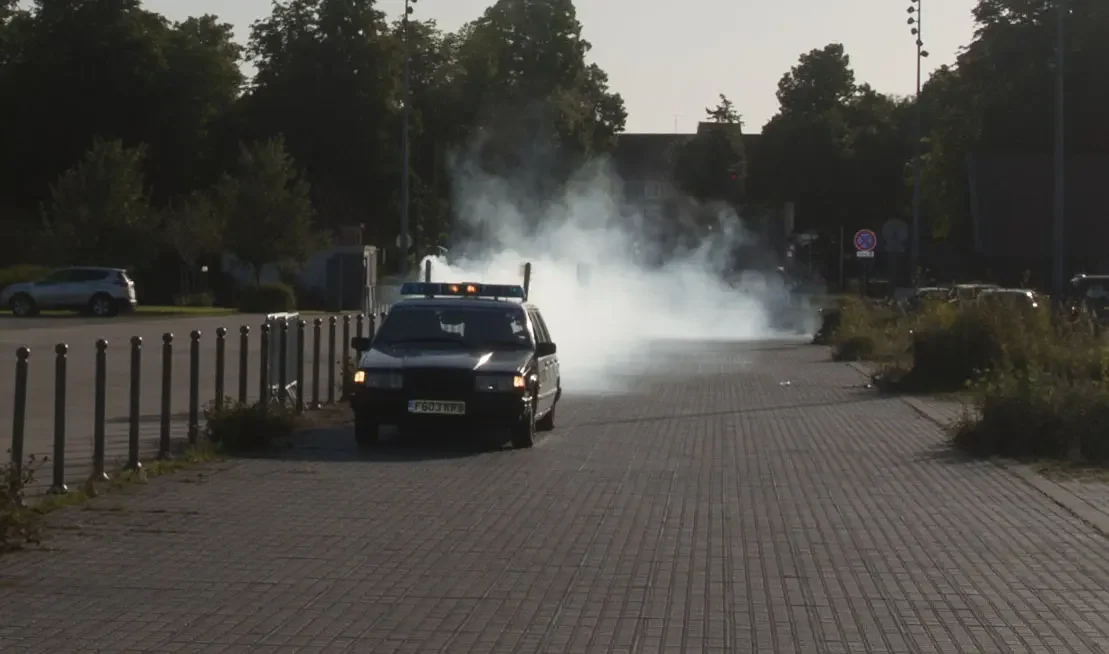 A black police car with a speaker on top is parked on the sidewalk as white smoke rises in the background.
