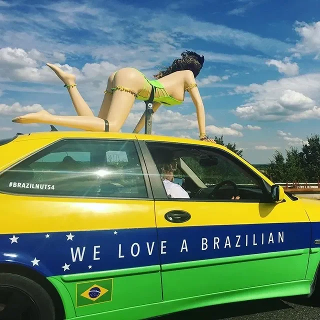A large inflatable female mannequin wearing a green bikini on top of a yellow and green taxi decorated with a Brazilian flag and the phrase 'WE LOVE A BRAZILIAN'.
