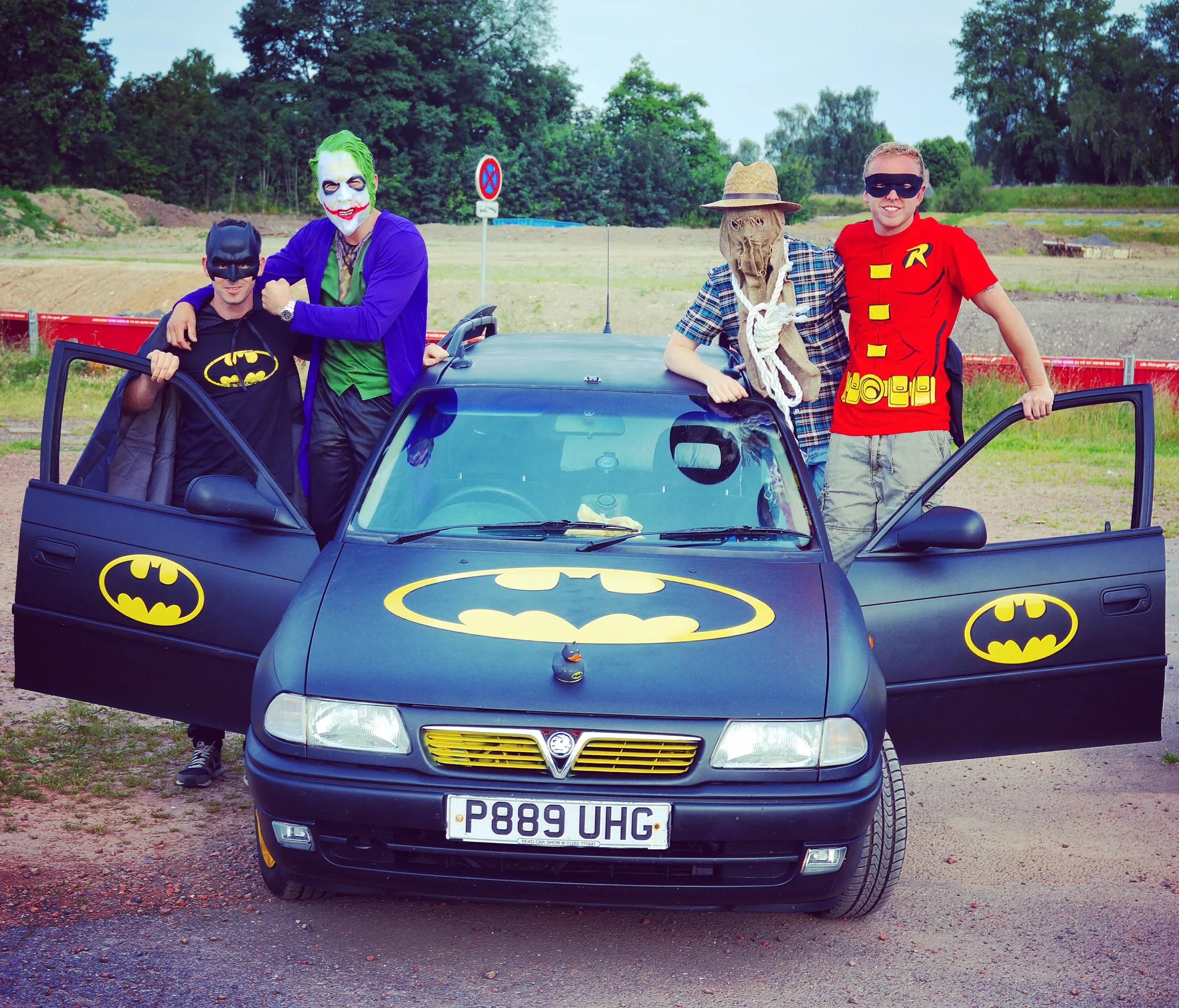 Four people in costume pose around a black Batman-themed car with Batman logos. One person is dressed as the Joker, another as Robin, and another as a scarecrow. The fourth person wears a black mask and Batman shirt. They are outdoors on a dirt and g