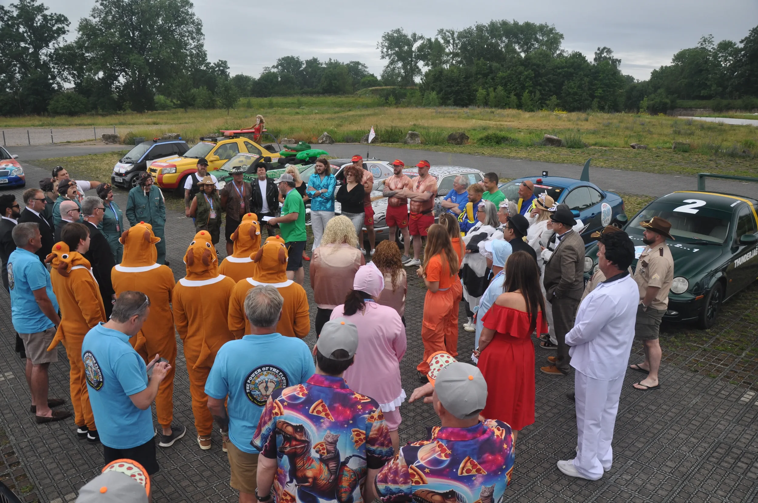 A gathering of people dressed in various costumes, including animals, characters, and casual attire, standing outdoors near several decorated cars resembling characters from 'The Amazing Race.'