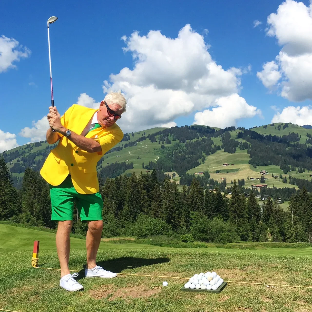A man wearing a yellow blazer, green shorts, white shoes, and sunglasses is preparing to hit a golf ball on a grassy golf course, with a mountainous landscape and blue sky with clouds in the background.