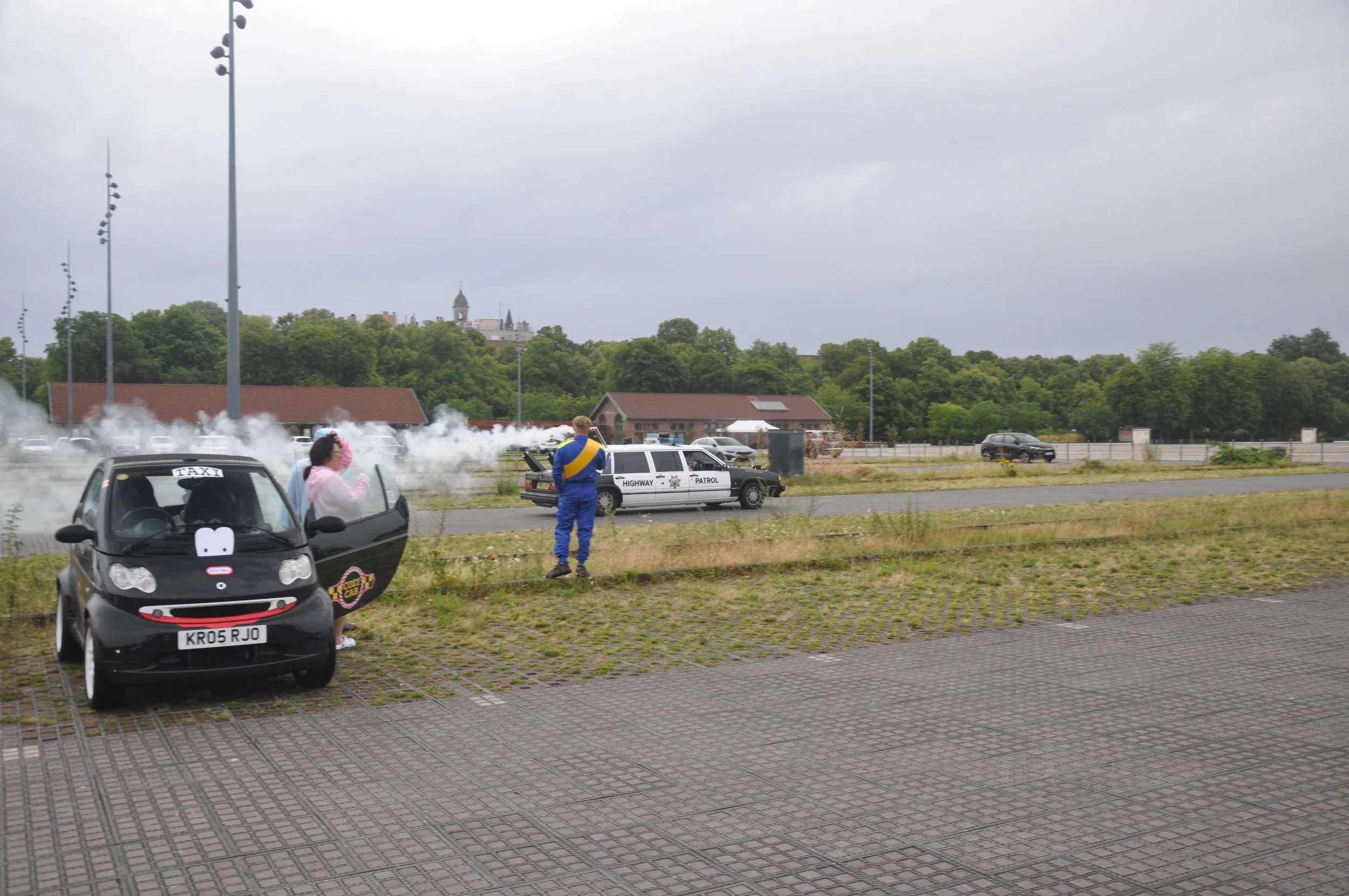 A scene with a small black taxi car parked on a paved area, a person in pink with a headscarf near it, a highway patrol police car in the background, and a man in blue overalls taking photos, with smoke or steam rising behind the police car. Green tr