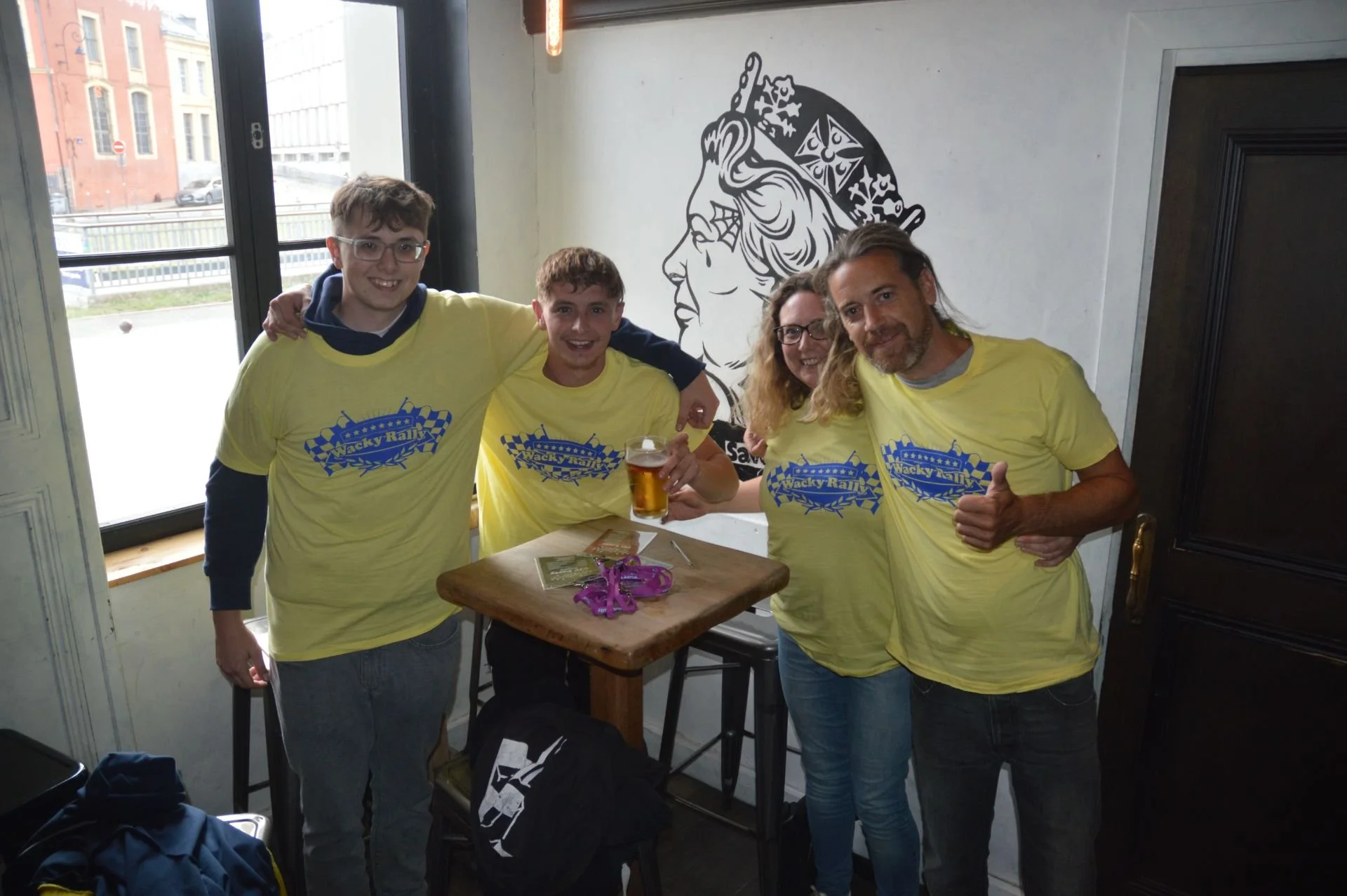 Group of four people smiling, wearing yellow 'Wacky Rally' shirts, in what looks like a bar or restaurant, with a window and a wall with a graphic of a woman's profile behind them. One person holds a glass of beer.