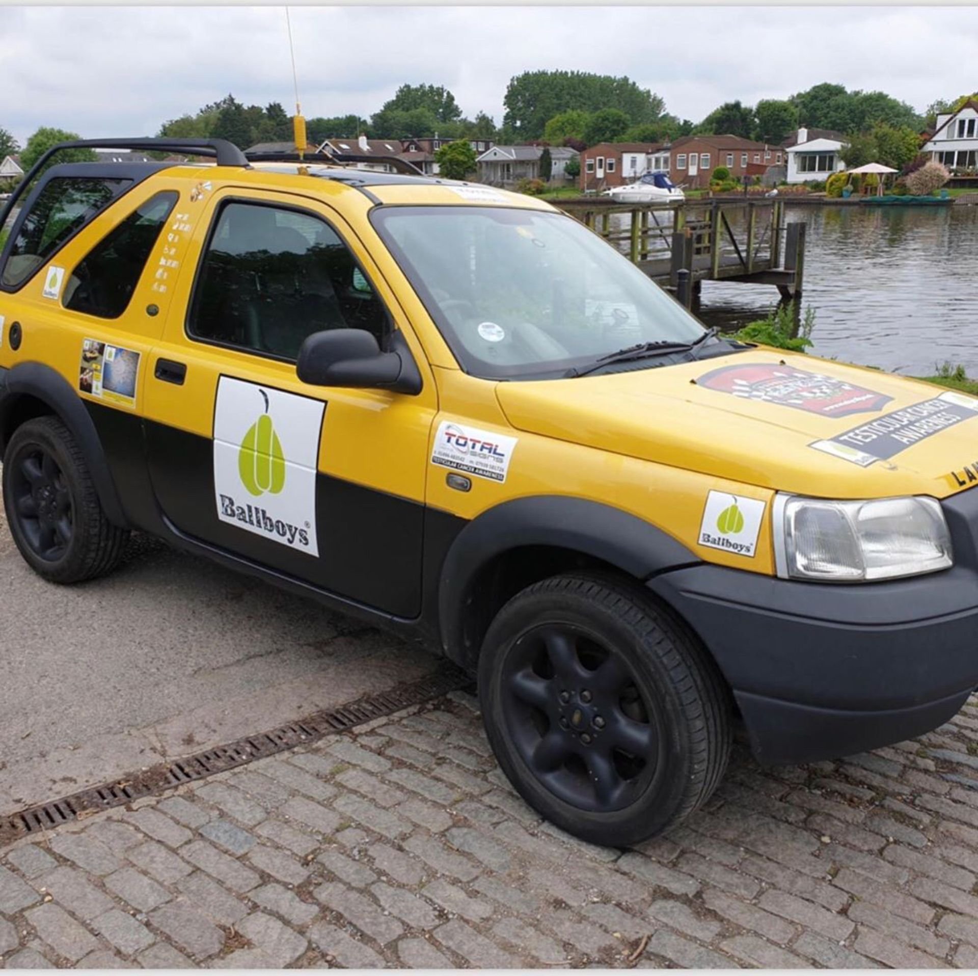 A yellow and black car parked on a cobblestone road next to a body of water with houses and trees in the background. The car has stickers and sponsor logos, including "Ballboys" and "TOTAL."
