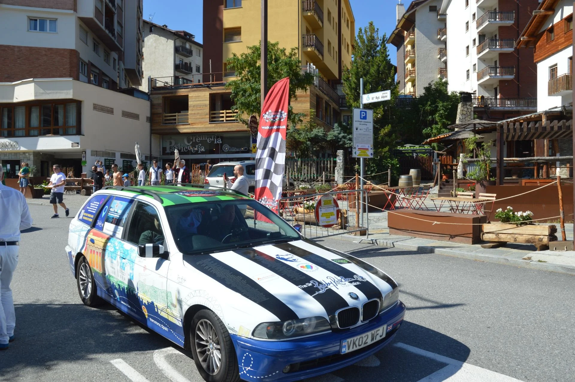 A street scene with a white and black decorated BMW car in the foreground, pedestrians walking on the sidewalk, colorful buildings, and a red flag with white annotations.
