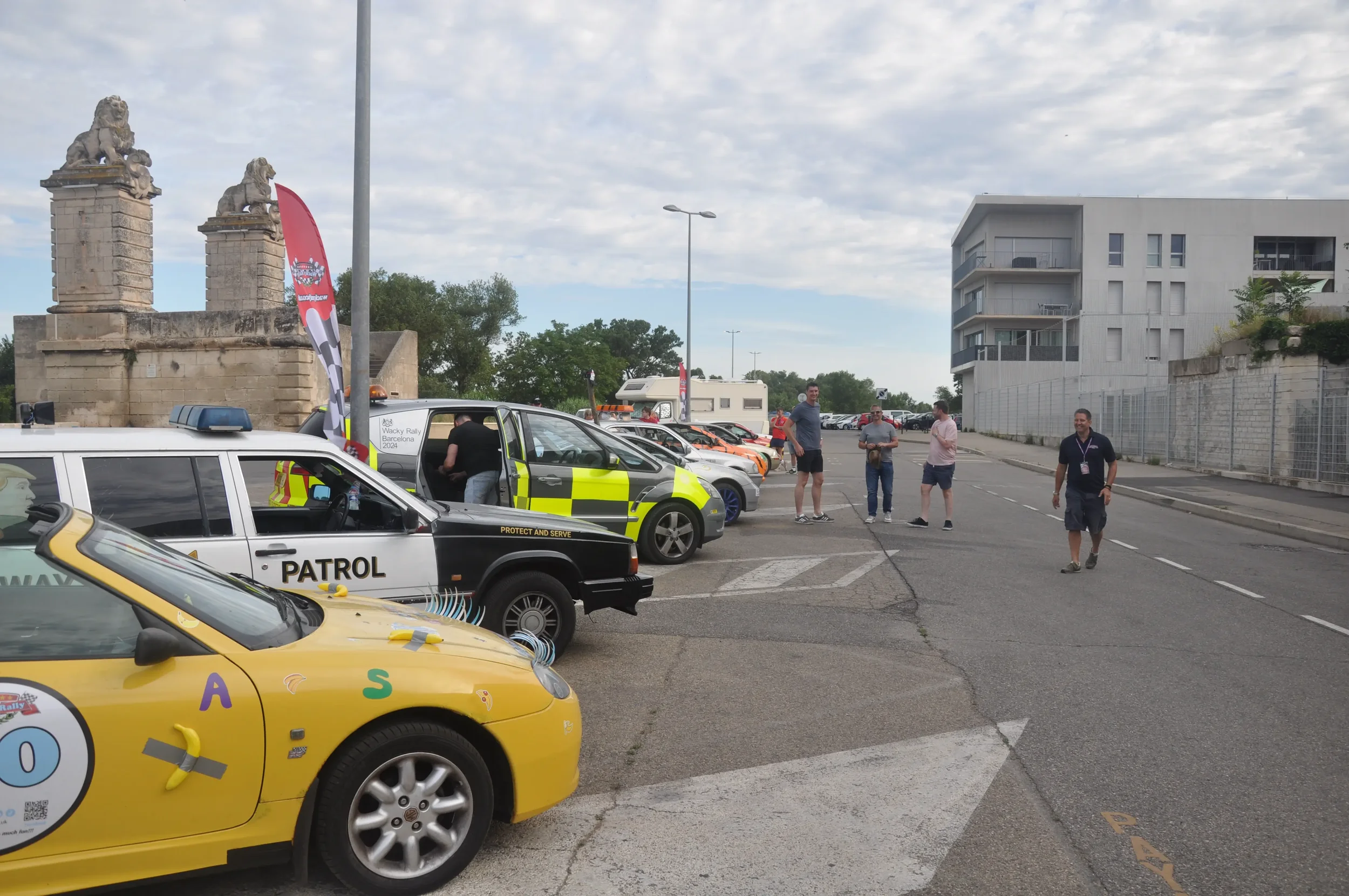 Lineup of rally cars parked along a street, with a few people walking and observing, and historic stone lions on structures in the background.