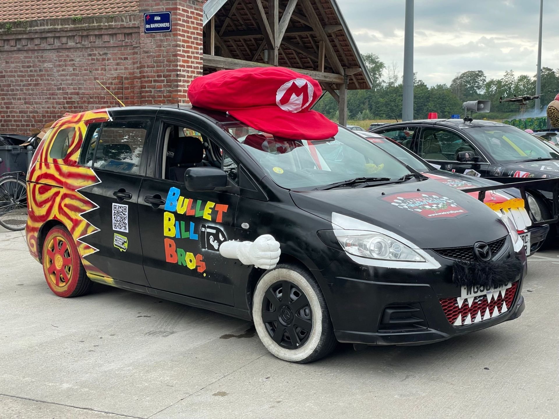 A black car decorated as Mario from the Super Mario video game franchise, with a large red Mario hat on the roof, a fabric beard, a mouth with sharp teeth painted on the front bumper, a white glove pointing forward on the side, and colorful lettering