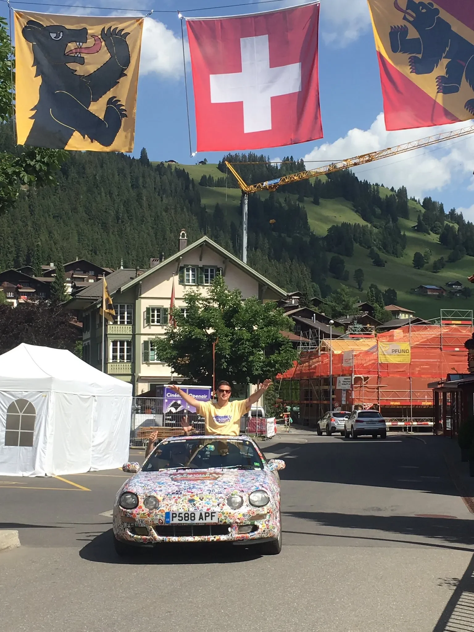 A person standing through the sunroof of a colorful, floral-patterned car with Swiss license plates, waving and smiling at a street festival in a Swiss town, with flags hanging overhead including a Swiss flag and two flags with a black lion on a yell