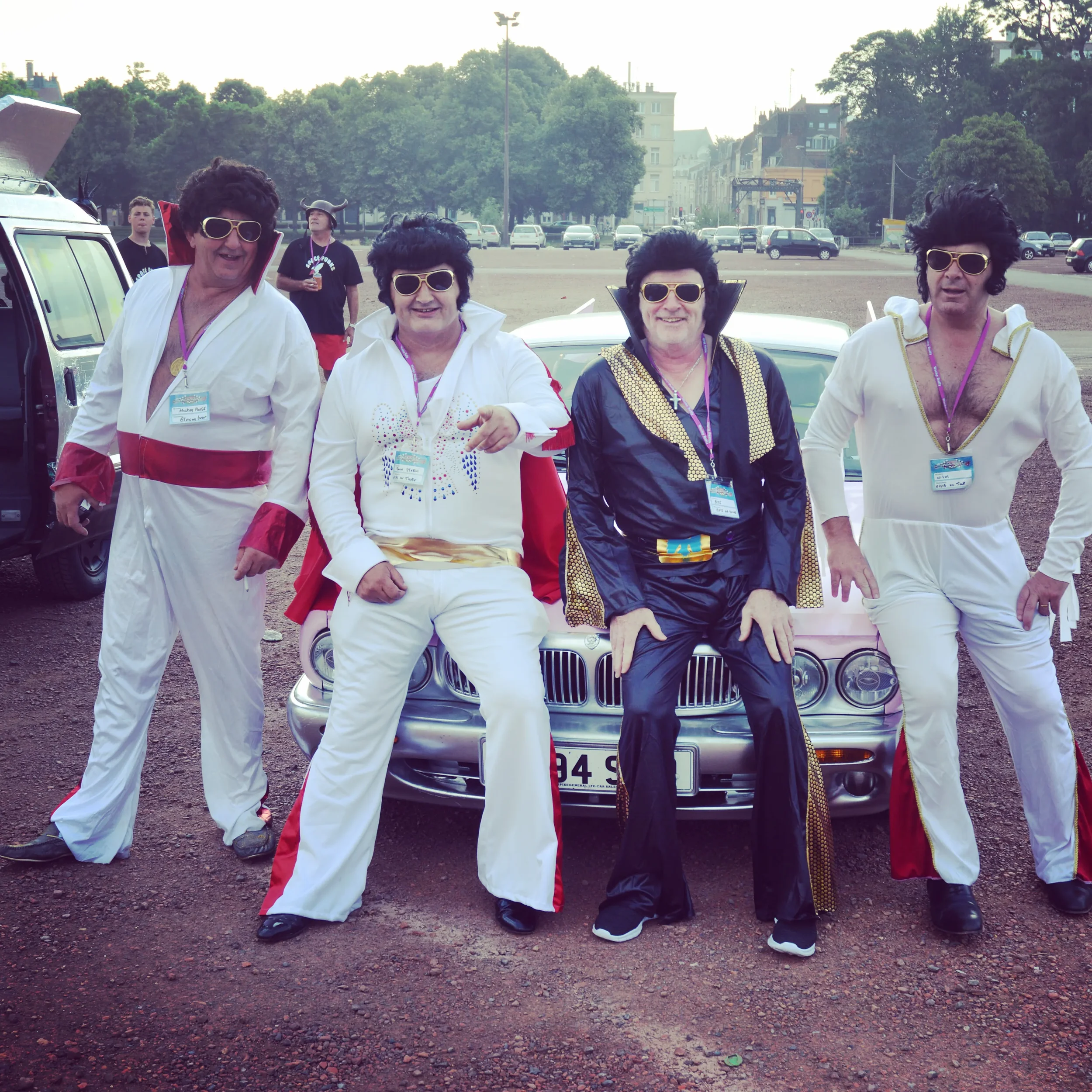 Four people dressed as Elvis Presley characters posing in front of a car with a parking lot and trees in the background.