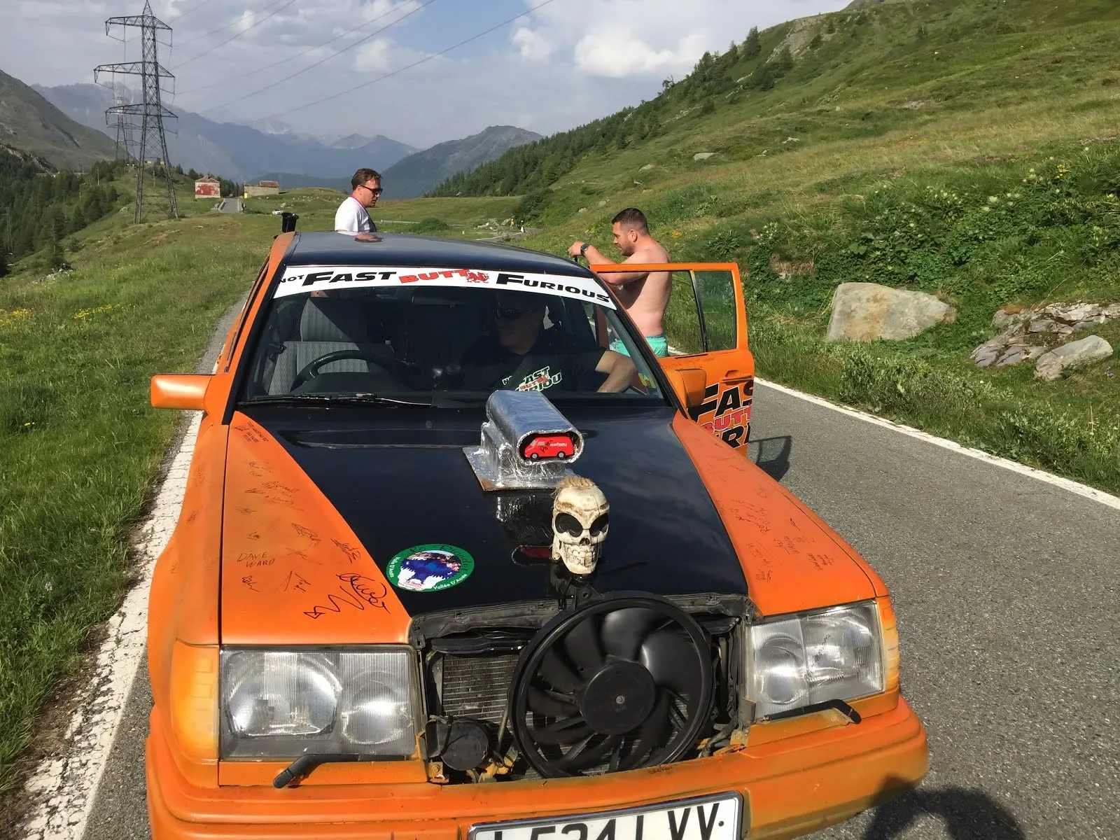 An orange vintage car with its front bumper removed, parked on a mountain road. The car has a black hood with signatures, a skull ornament, and a model engine on the hood. There are three men around the car, one inside, one shirtless and standing nea