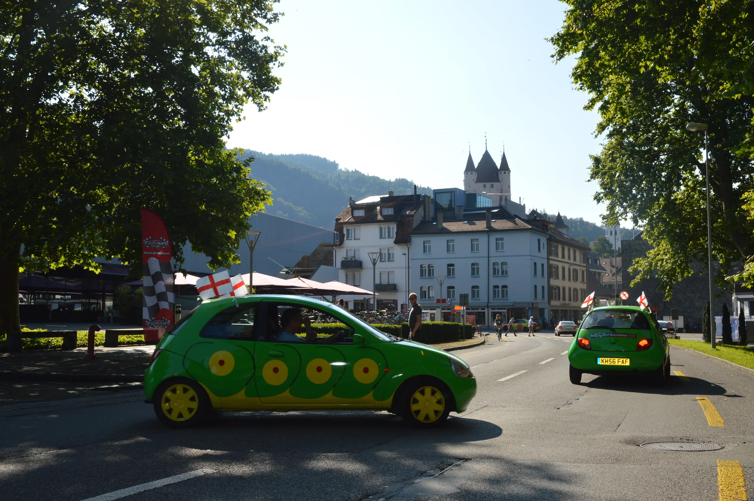 Two small green cars decorated with England and patriotic flags on a street in a city with a castle on a hill in the background, with people walking and a person standing near one car.